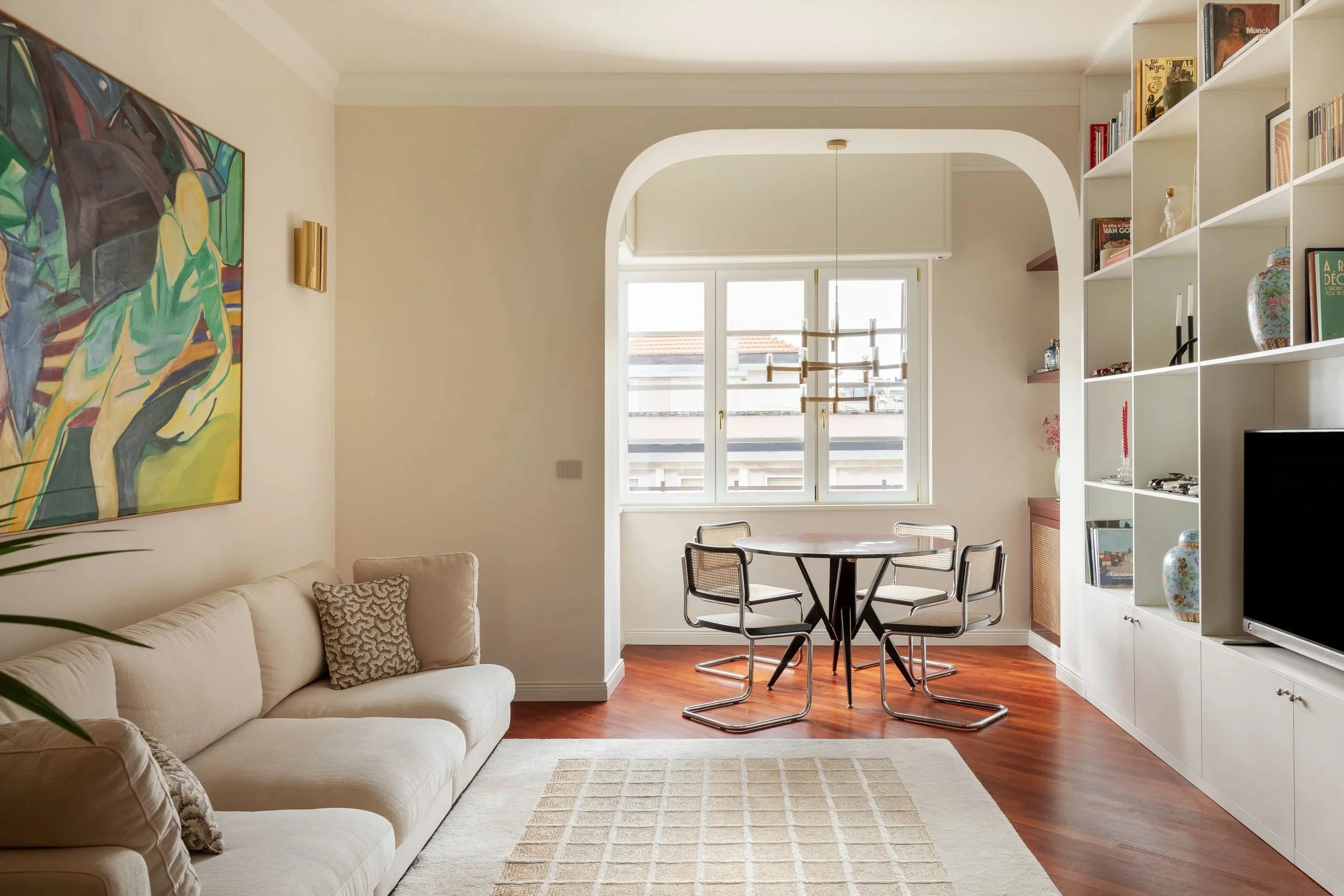 Interno di un soggiorno con un divano beige, pareti chiare, quadro astratto e libreria bianca con oggetti decorativi, grande finestra con vista sul cielo e mobili moderni.