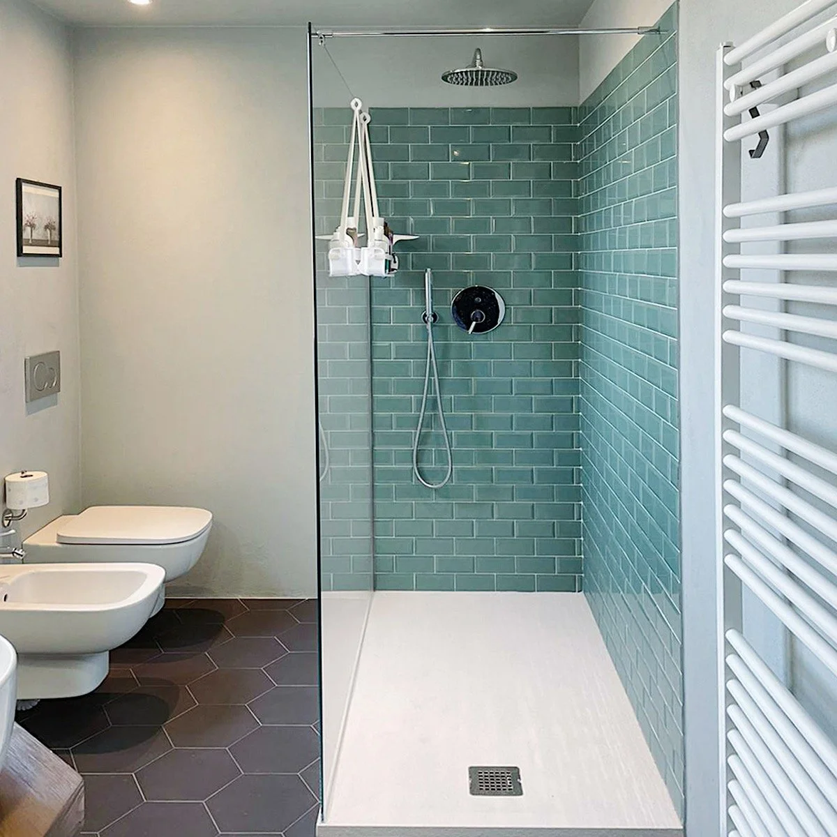 Bagno con doccia a filo pavimento, piastrelle verdi in stile subway, lavabo e WC sui lati, termosifone bianco a barre verticale, pareti bianche, pavimento a piastrelle esagonali marroni.