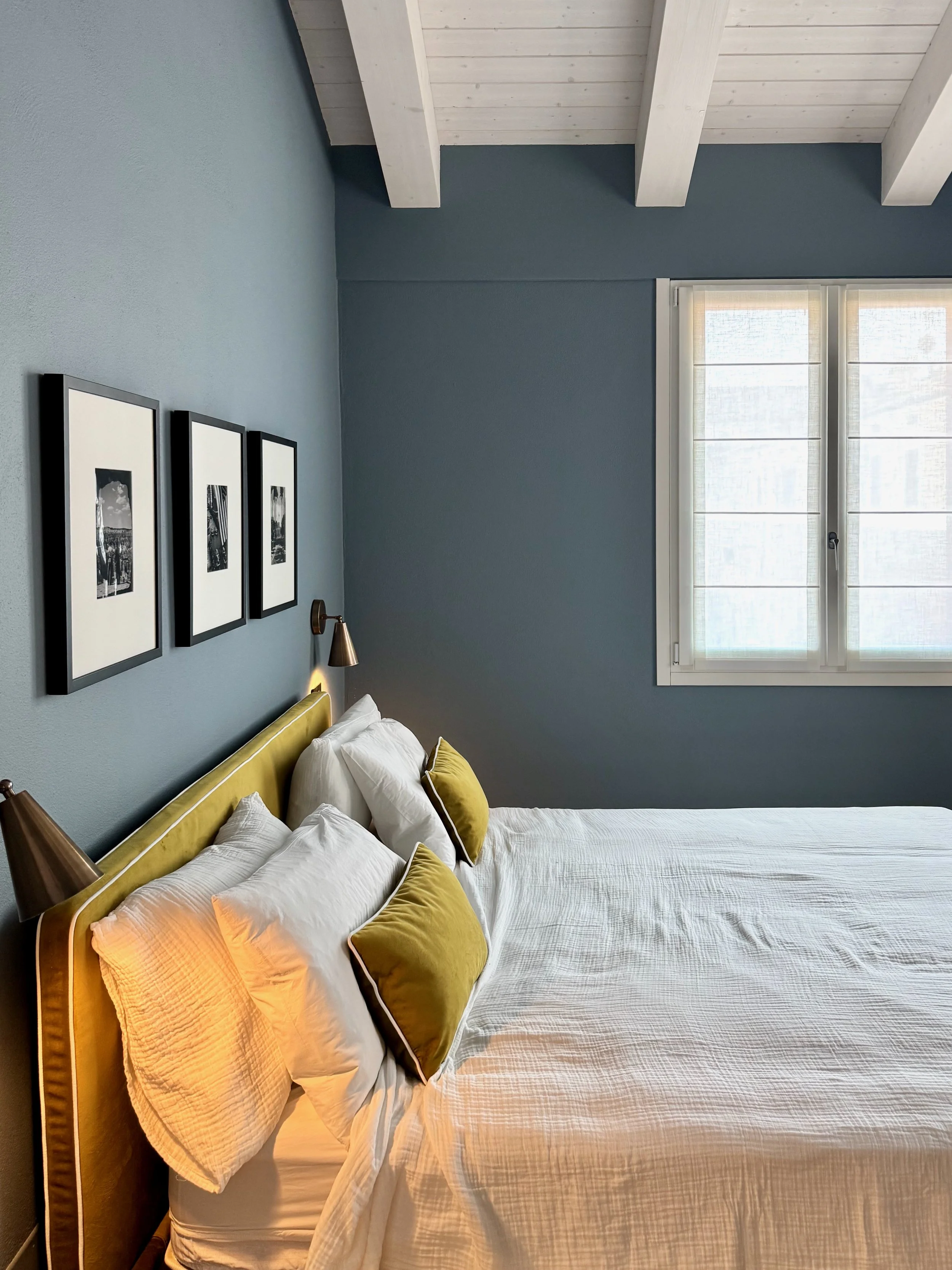 Una camera da letto con pareti blu, un letto con lenzuola bianche e cuscini gialli, un quadro con tre immagini nere e bianche appeso alla parete e una finestra con tende chiare.