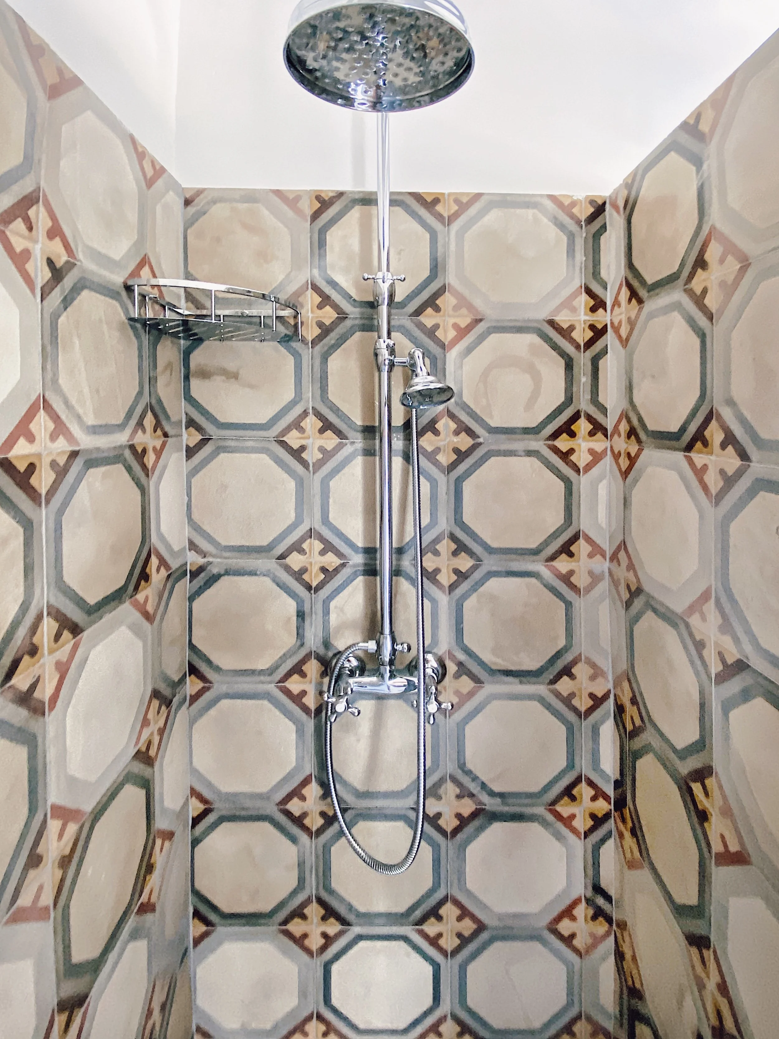 Piastrelle decorate con pattern geometrico in tonalità di beige, verde e marrone in una doccia con soffione rotondo, maniglia e doccetta a mano.