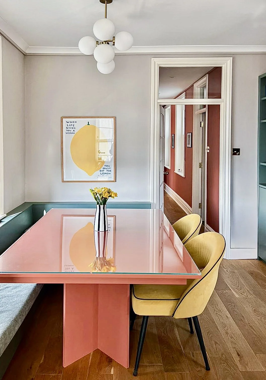 Sala da pranzo con tavolo rosa, sedia gialla, vaso di fiori gialli, quadro con limone, lampadario bianco e muro bianco, ingresso con porte bianche e pareti rosse.