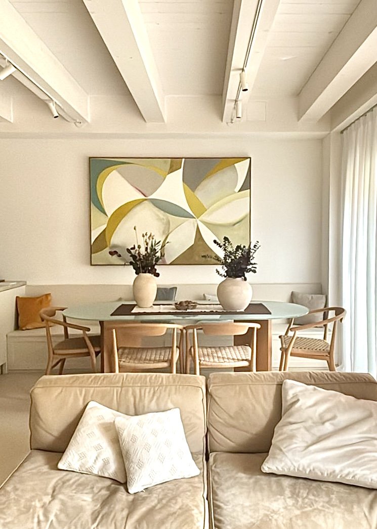 Soggiorno con divano beige, cuscini bianchi, tavolo da pranzo ovale con sedie di legno, due vasi con fiori e un quadro astratto sulla parete