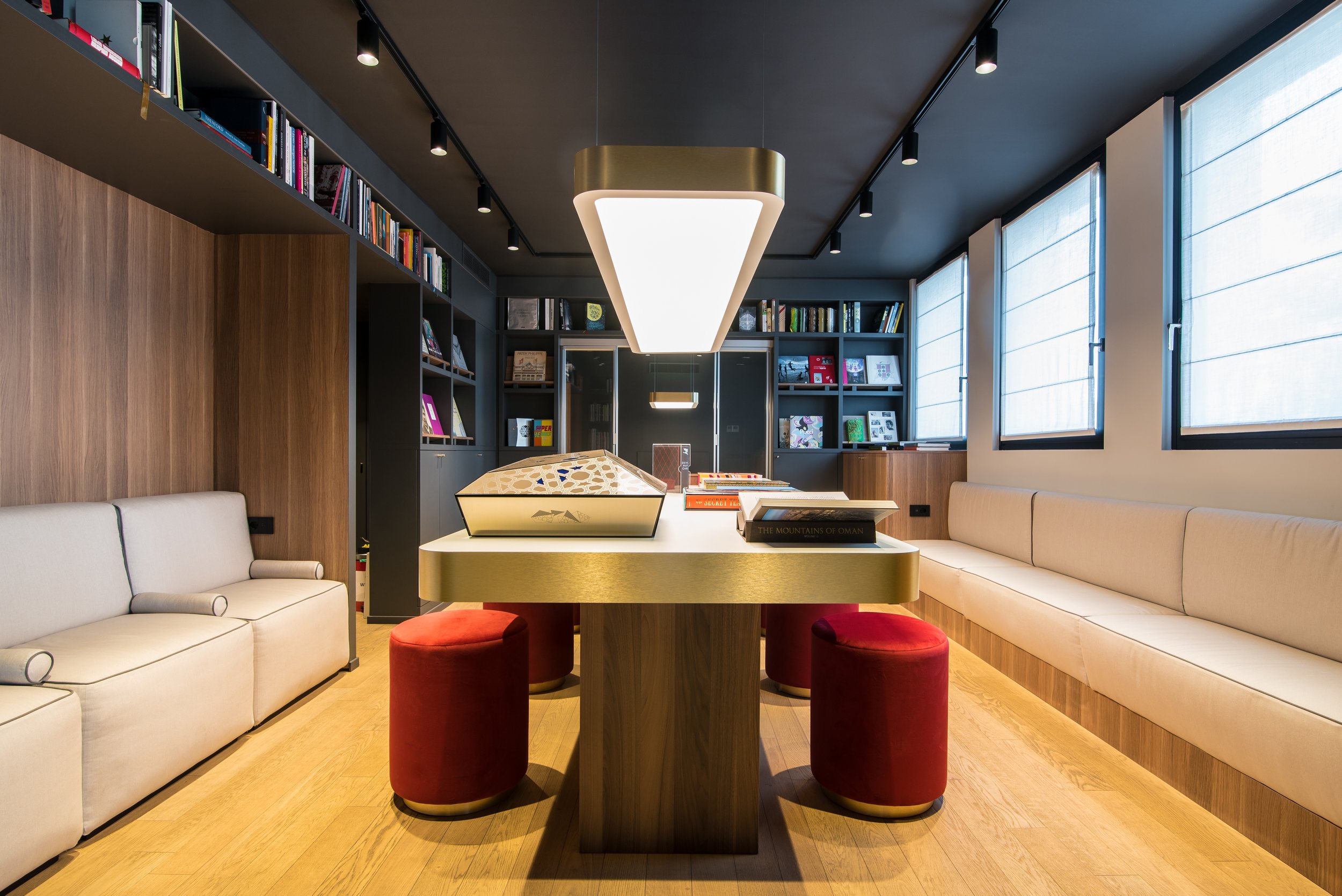 Sala con tavolo centrale, sedie rosse, librerie sullo sfondo, grandi finestra, illuminazione moderna.