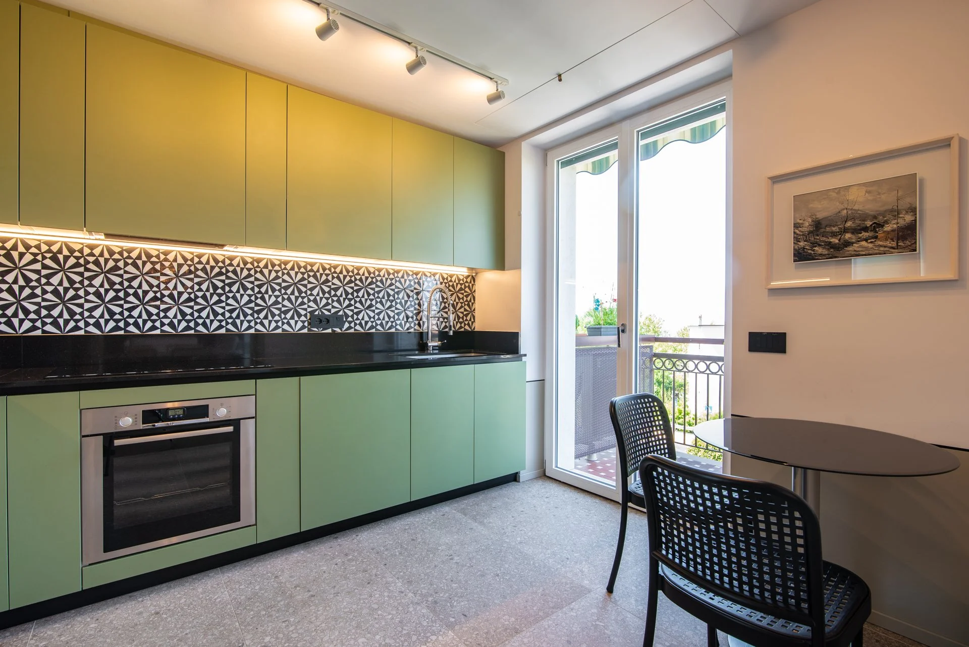 Cucina moderna con mobili verde chiaro, schermo a piastrelle geometriche nero e bianco, balcone visibile attraverso la porta finestra, con tavolo nero e due sedie.