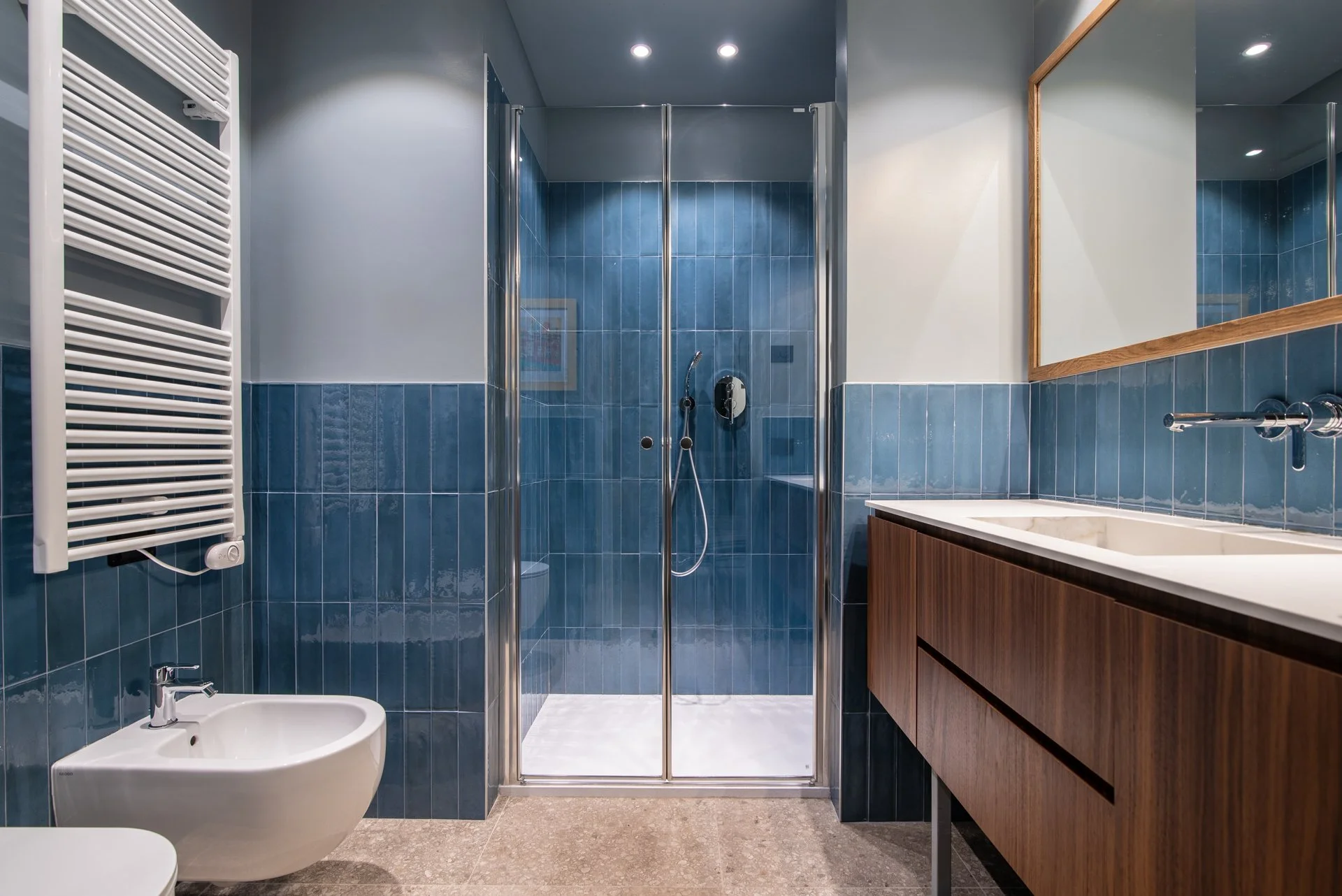 Bagno moderno con pareti rivestite di piastrelle blu e pavimento in pietra, con lavabo in legno, grande specchio e cabina doccia in vetro.