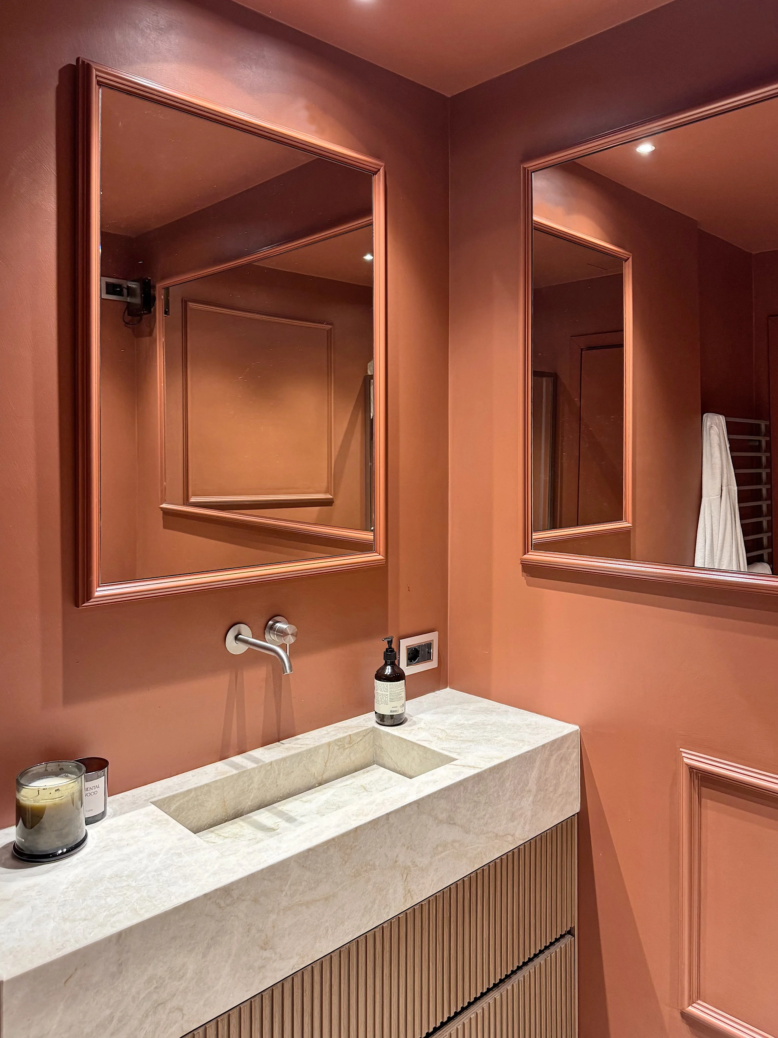 Specchio rotondo sopra lavabo in marmo bianco, pareti color terracotta, bagno con specchi rettangolari sulle pareti, tenda bianca appesa, accessori per il bagno come sapone e candele.