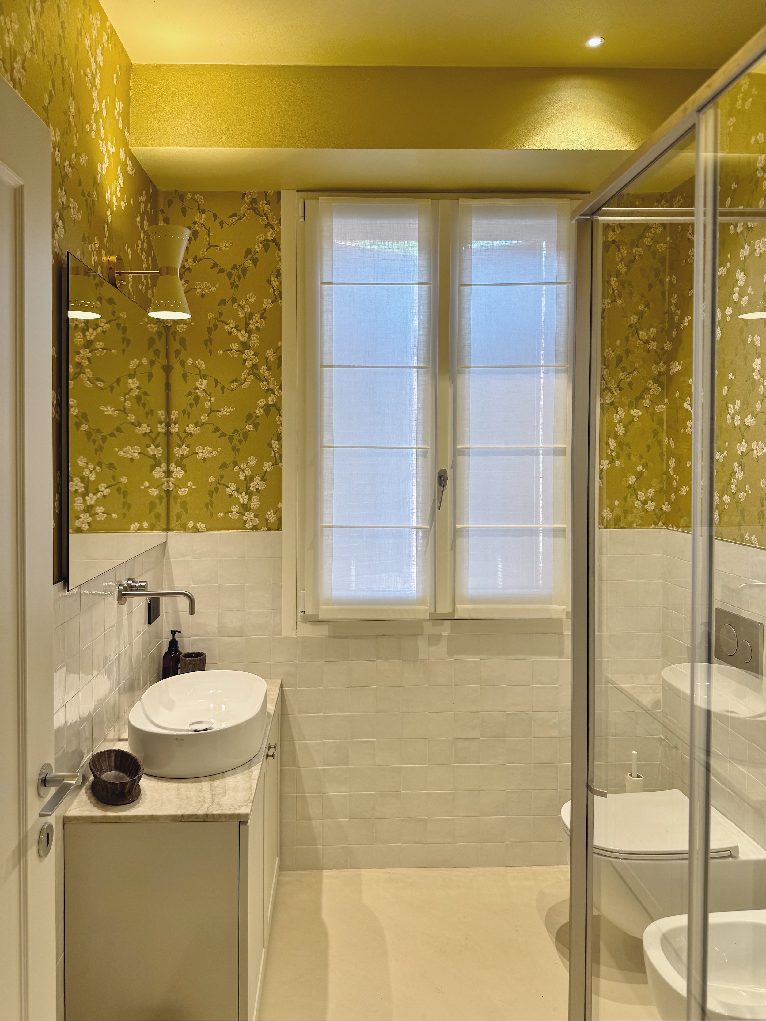 Foto di un bagno con pareti tappezzate di carta da parati gialla con fiori bianchi e rami verdi. Fenetre centrale con tende chiare. Sul lato sinistro, lavabo bianco con mensola in marmo, al di sopra una specchiera e una luce da parete. Sul lato destr