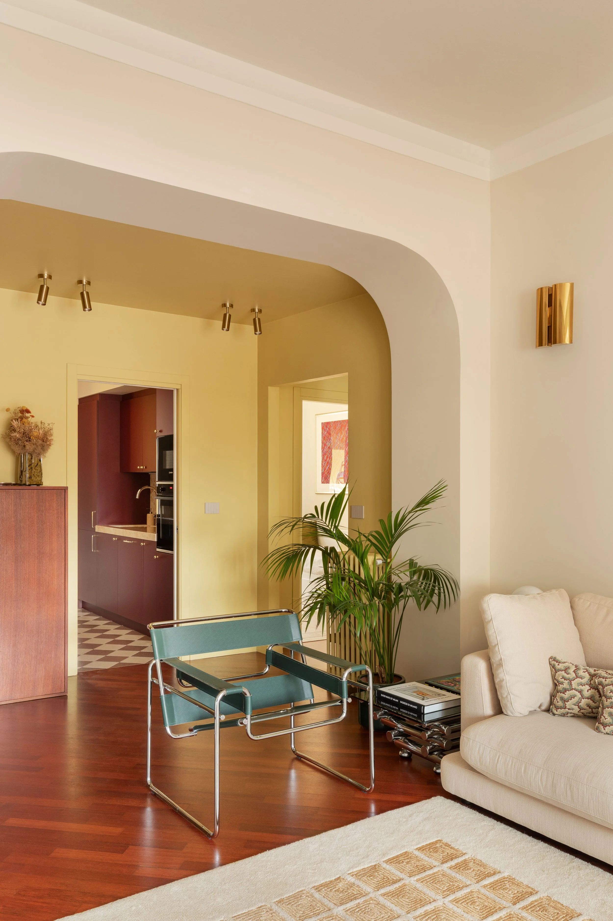Interno di soggiorno con divano bianco, piante verdi, tavolino con riviste, sedia moderna in acciaio e tessuto blu, open space con pareti gialle e parete con quadri, luci da soffitto e muro in ceramica con scacchi.