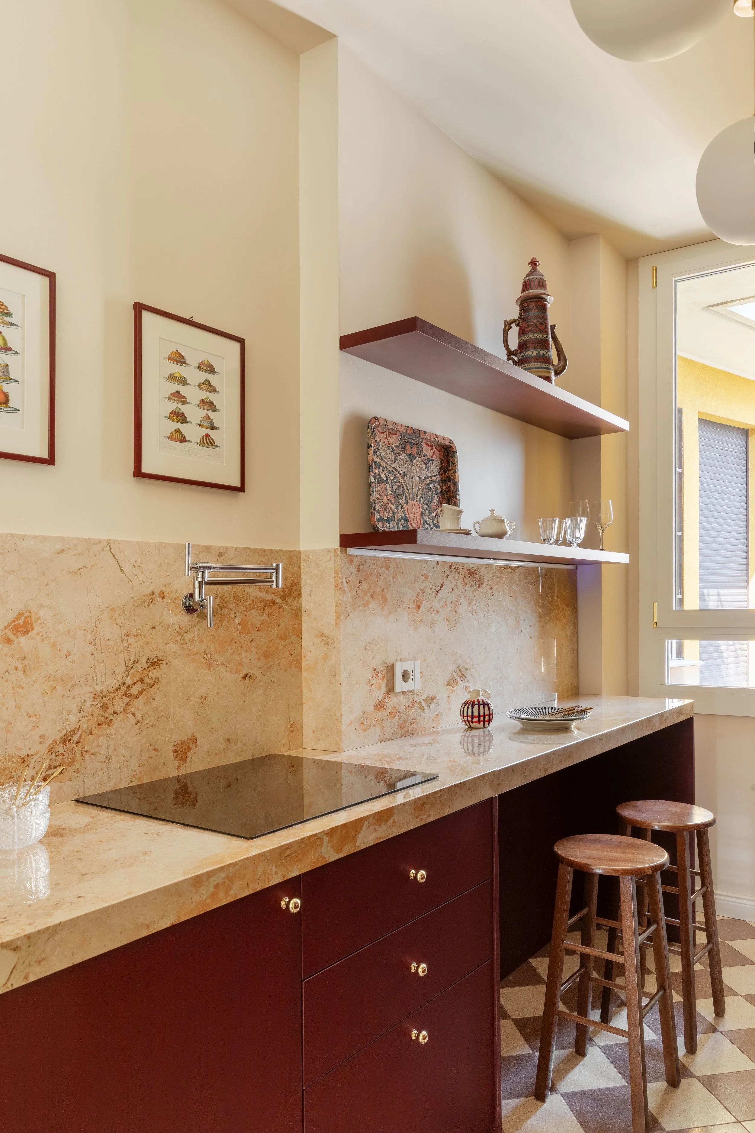 Cucina con bancone in marmo, tre sgabelli in legno, quadri con dolci alle pareti, mensole con ornamenti e bicchieri, porta finestra che dà su un balcone, pavimento a scacchi.