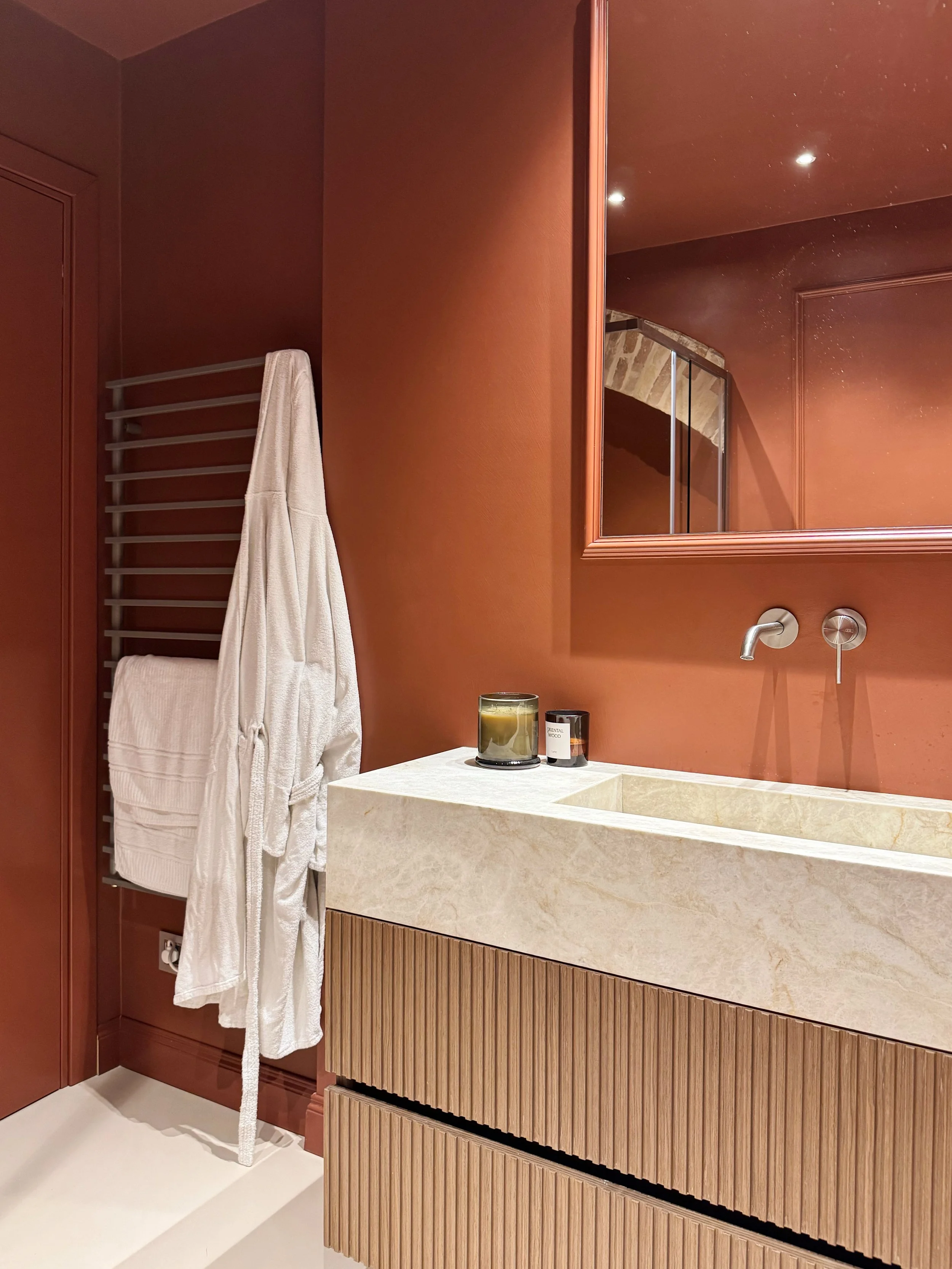 Specchio sopra un lavabo di pietra con mobili in legno, pareti color mattone, asciugamani bianchi appesi a un supporto a parete, candele e valvola del rubinetto visibili.