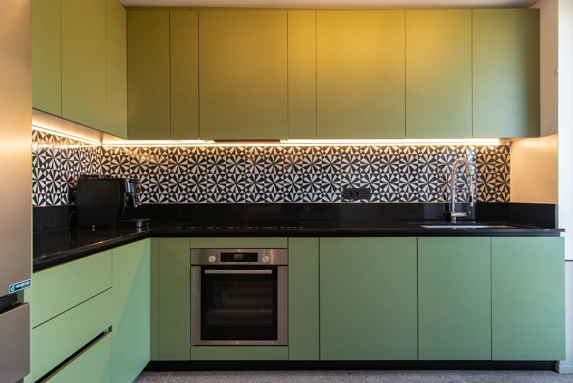 Cucina moderna con ante verde, piano di lavoro nero e rivestimento a piastrelle con motivo geometrico in bianco e nero, include forno, lavello con rubinetto e macchina da caffè.