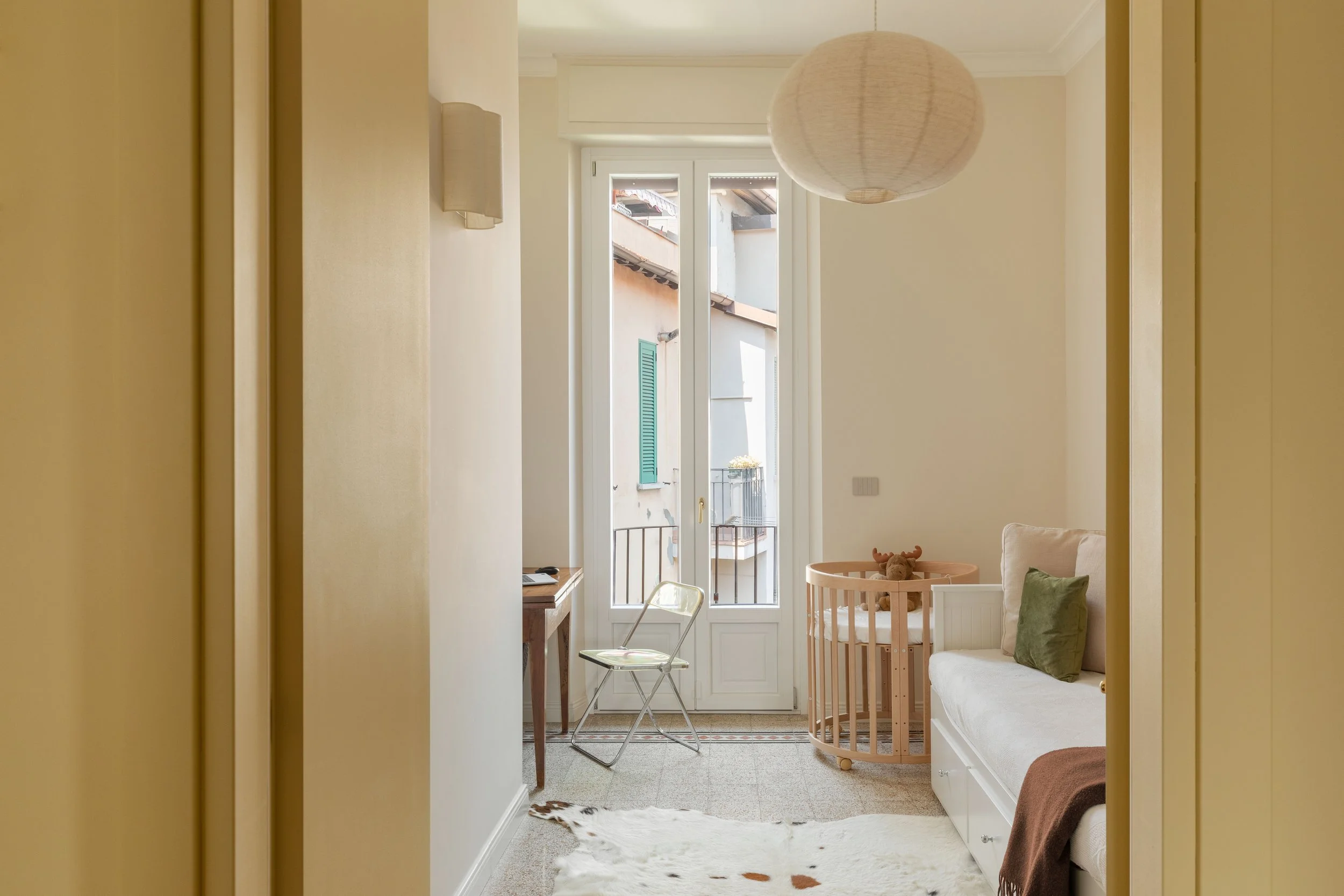 Interno di una stanza con pareti di colore beige, una finestra con persiane verdi e una porta di vetro. È presente un divano con cuscini, un tavolo rotondo con un giocattolo e una sedia trasparente. Sul pavimento c'è un tappeto bianco con macchie di 