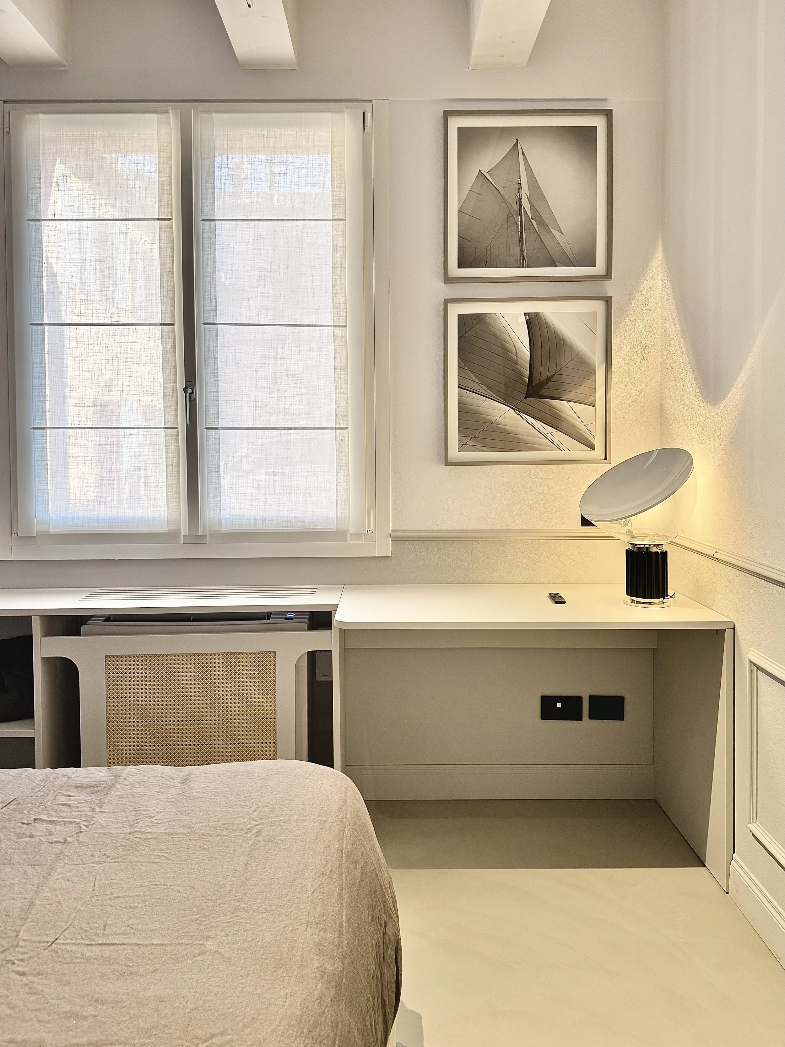 Interno di una stanza con una finestra con tende, un quadro con immagini di vele di barche, una lampada da tavolo moderna e un mobile bianco con prese di corrente.