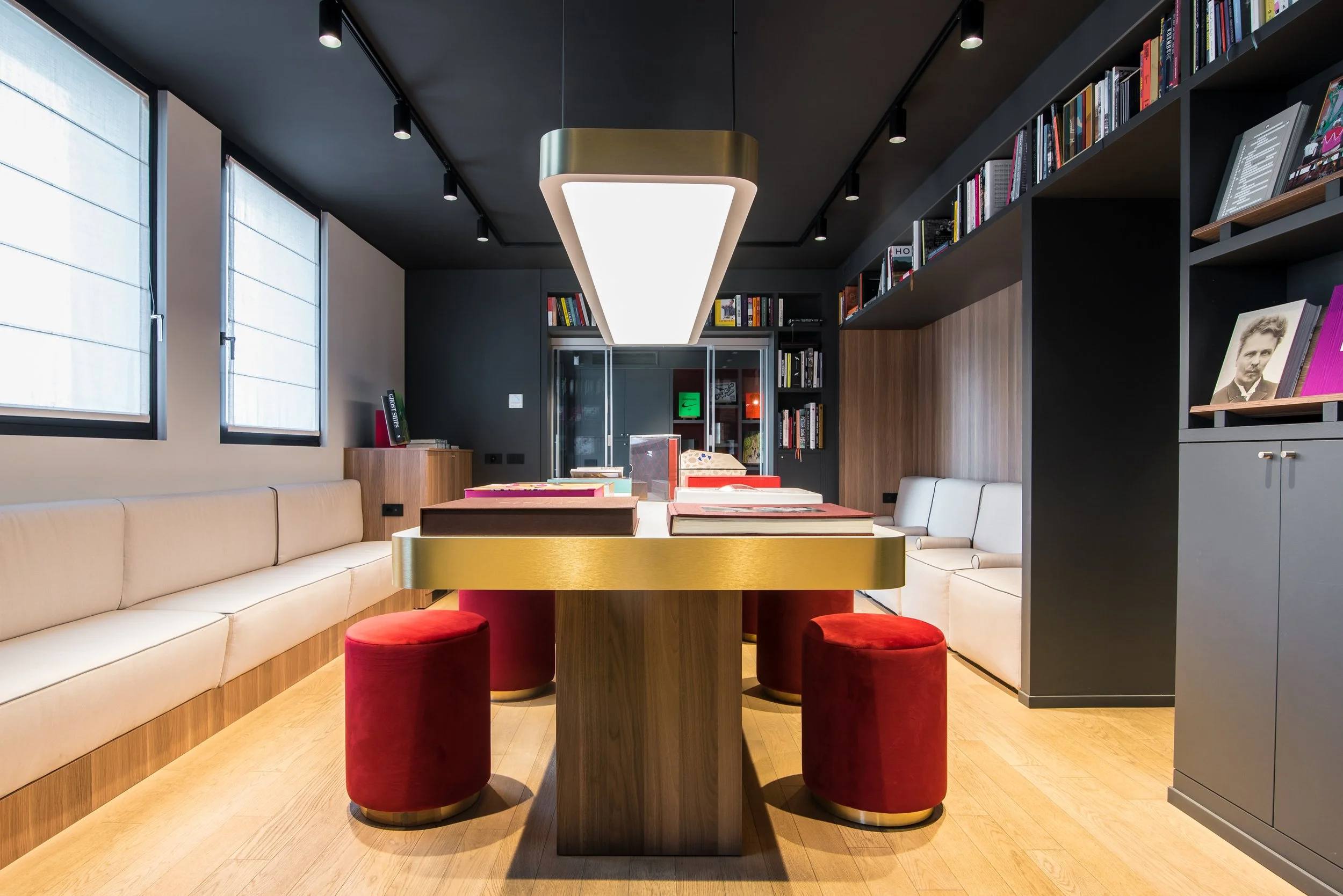 Una stanza con scaffali pieni di libri, un tavolo centrale con libri su di esso, sedie rosse e un divano beige lungo le pareti, una luce sospesa al soffitto e due grandi finestre con tende chiare.