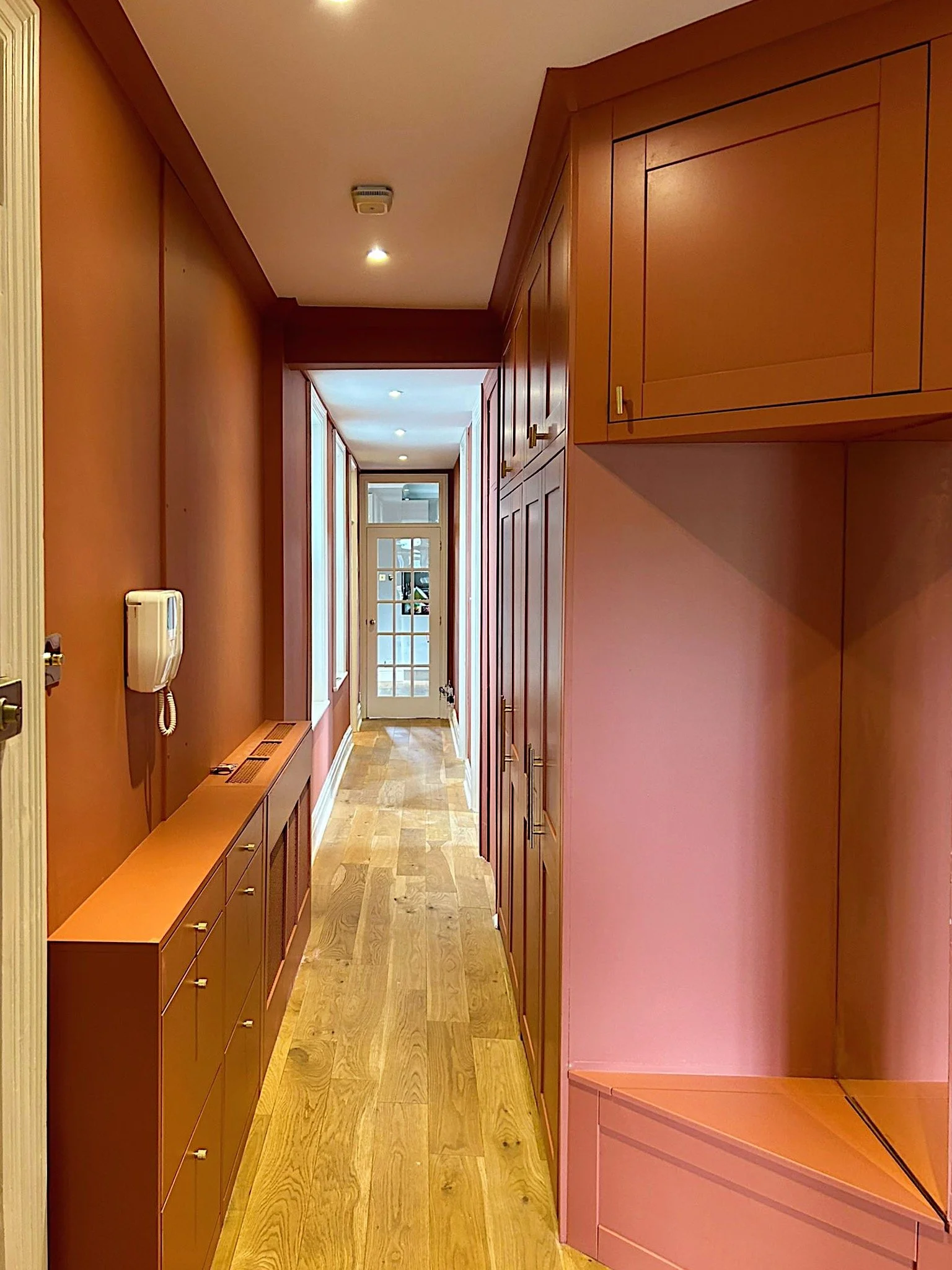 Corridor con pareti rosa e mobili in legno, porta finestra in fondo, pannello di controllo all'ingresso.