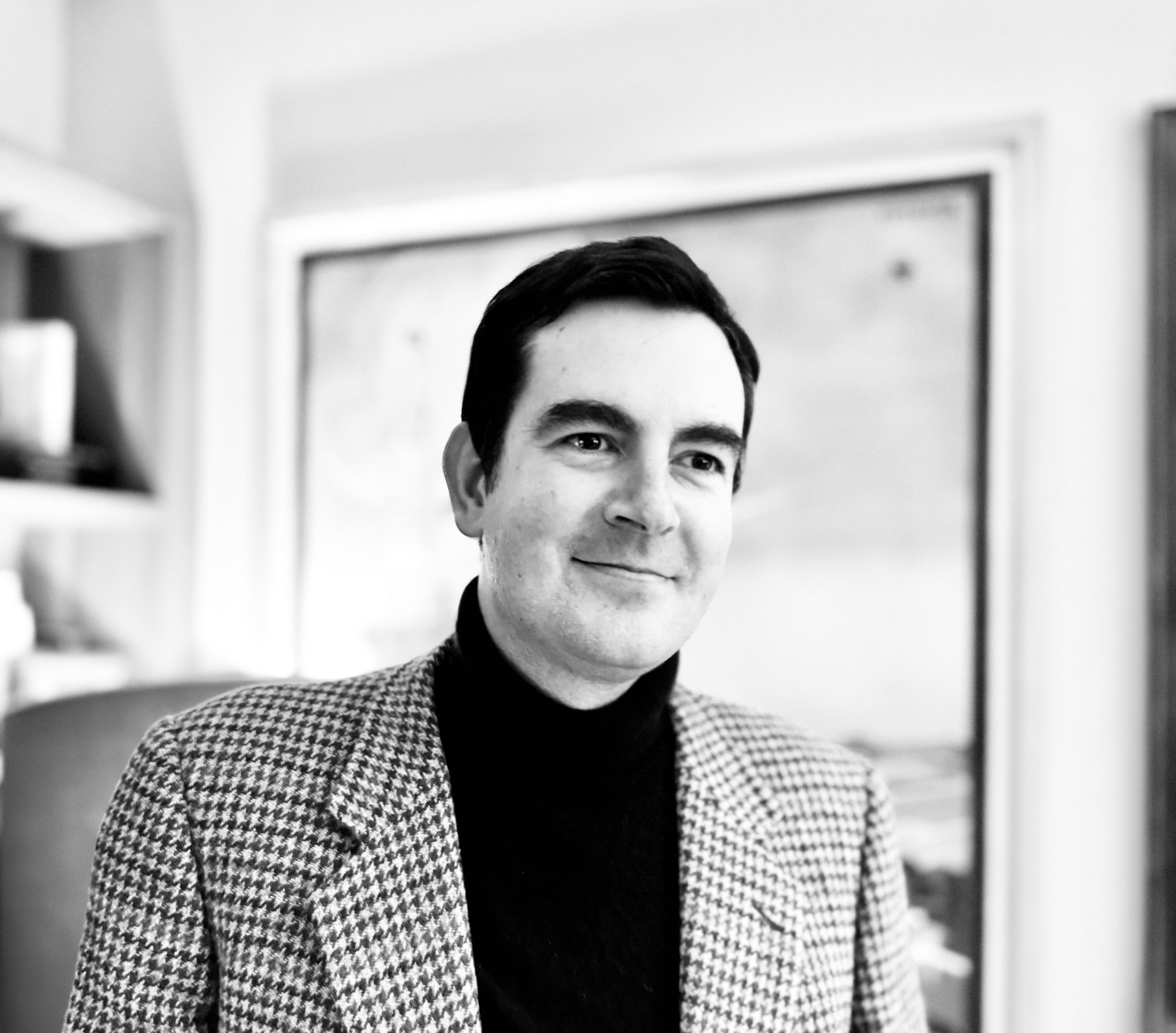 Un uomo sorridente con abbigliamento elegante, sfondo di un ufficio o stanza interna.