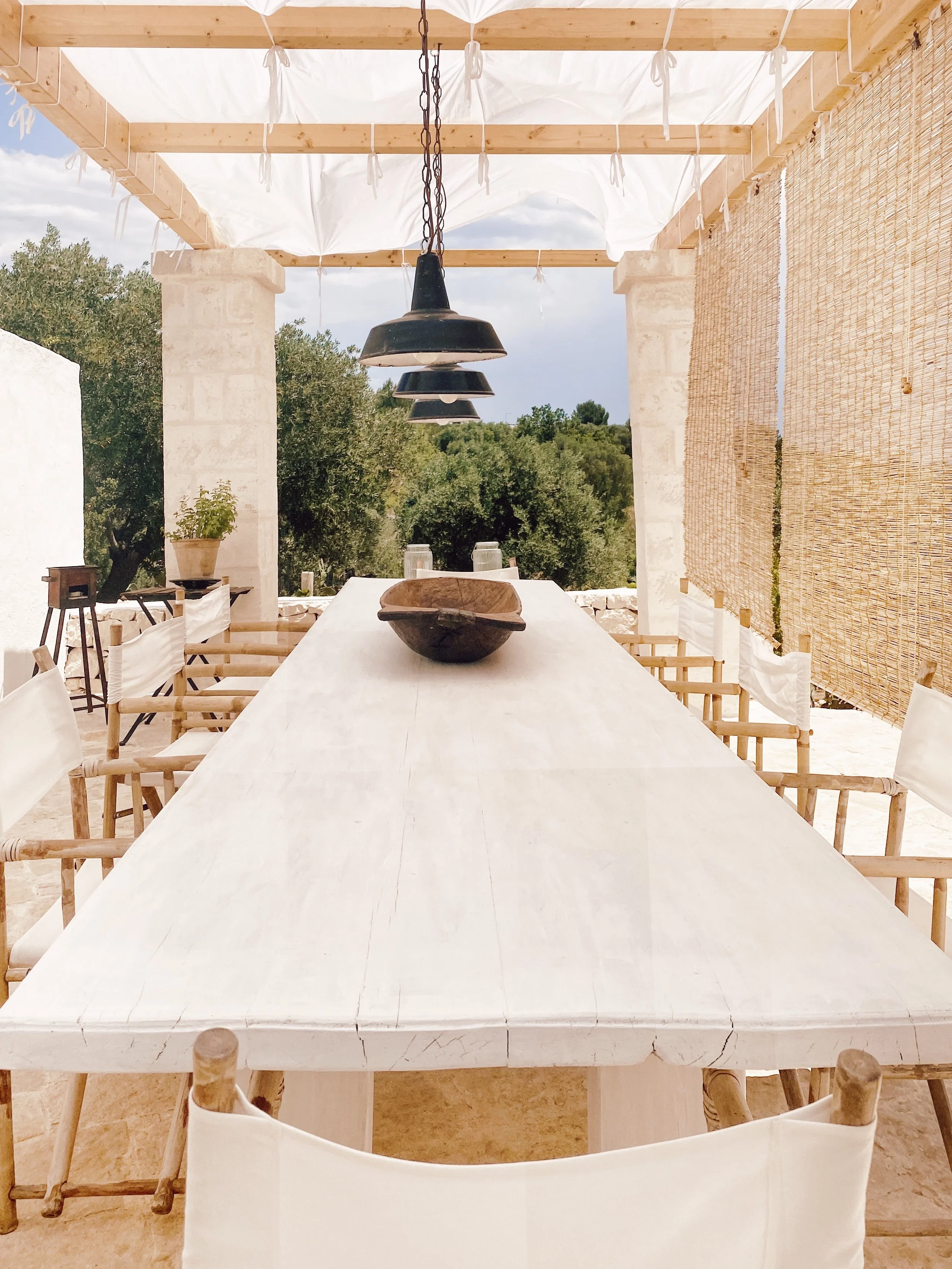 Tavolo da pranzo all'aperto con sedie di legno e schienali di tessuto bianco, pendant neri sopra, sfondo con natura e cielo.