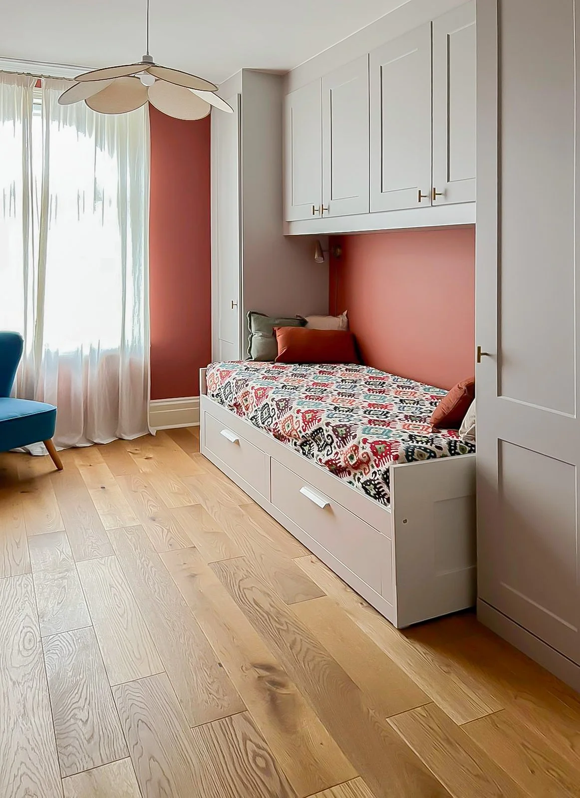 Camera con letto a castello bianco e testiera con cuscini, parete rosa e armadiature bianche. Finestra con tende leggere e sedie blu.