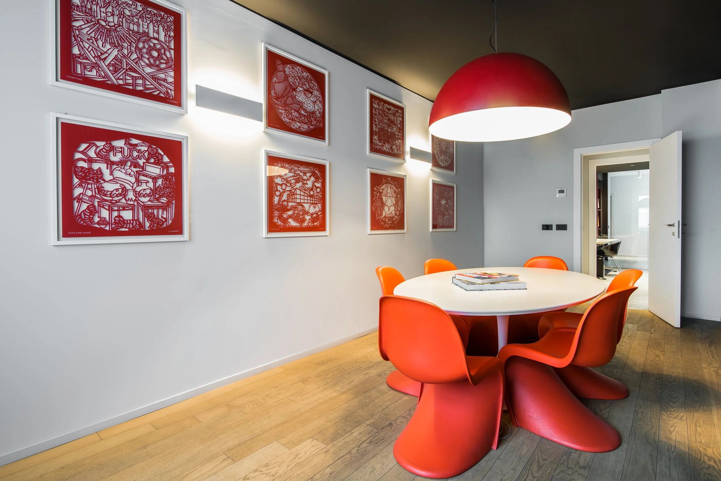 Sala da pranzo con tavolo bianco e sedie arancioni, parete decorata con quadri rossi, grande lampada rossa sopra, pavimento in legno, porta aperta su un'altra stanza.