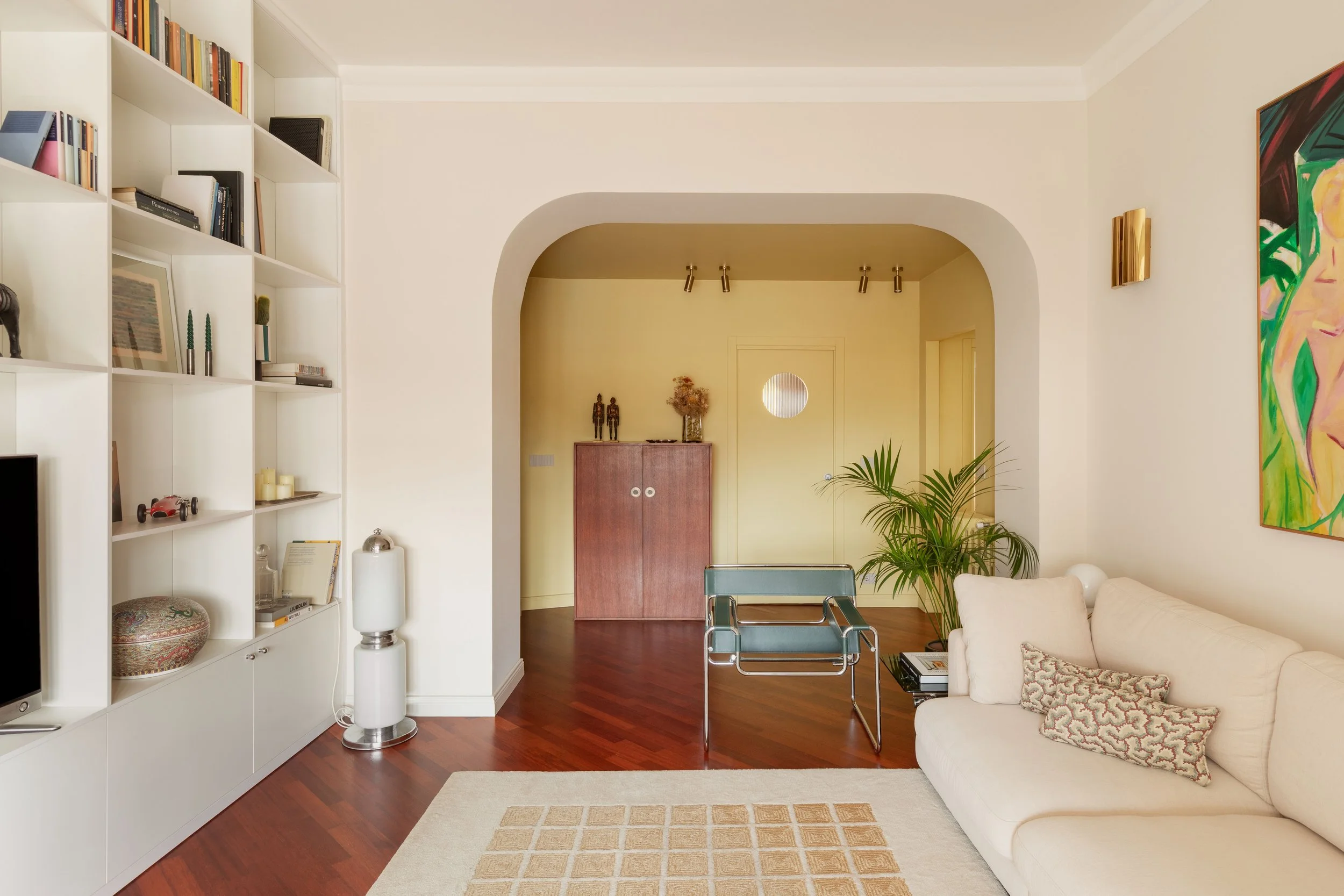Interior di un soggiorno con libreria bianca a sinistra, divano bianco a destra, pavimento in legno, dipinti colorati alle pareti, piante verdi e arredamento moderno.