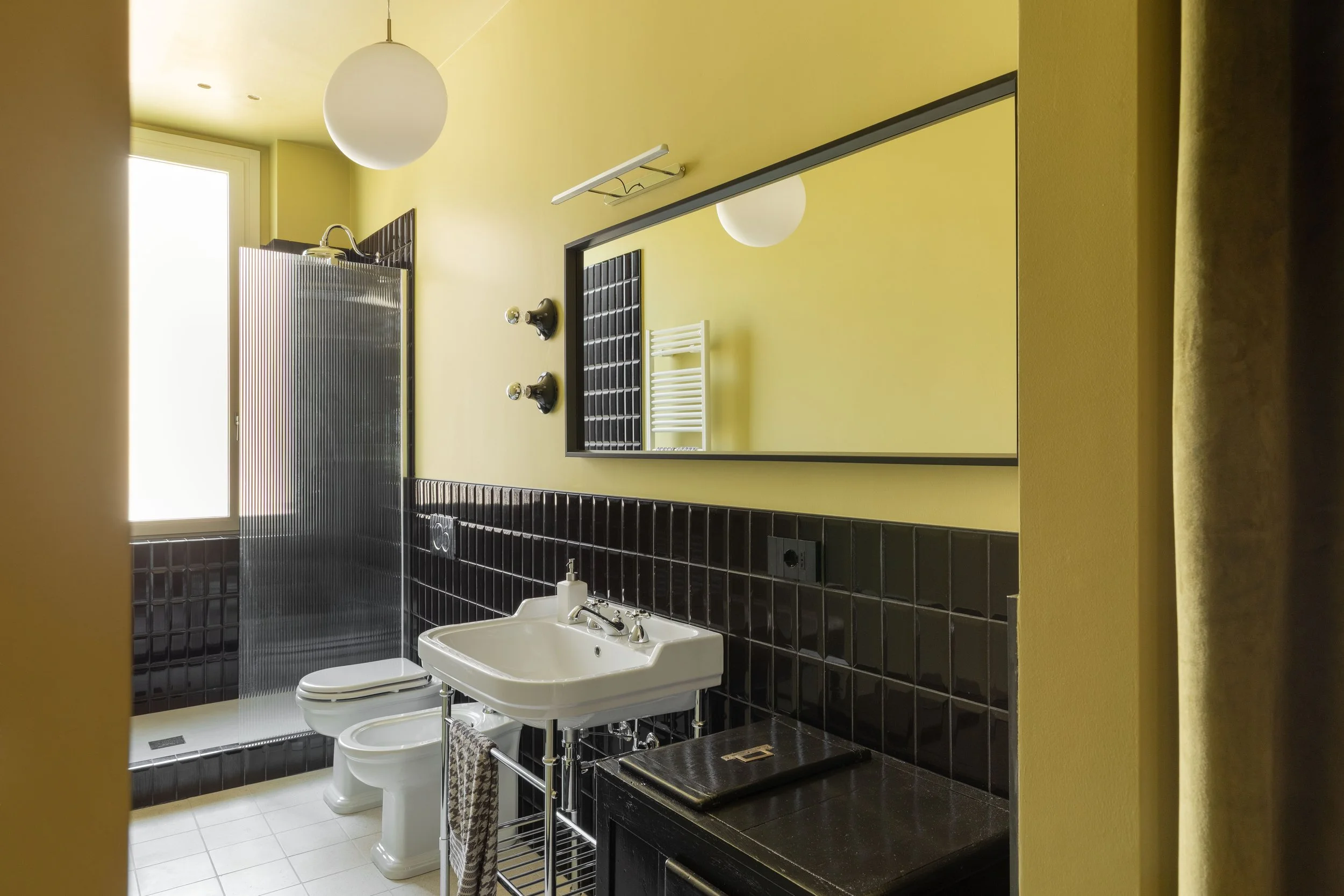 Bagno con pareti gialle e piastrelle nere, lavabo bianco, specchio, radiatore e lampada a soffitto