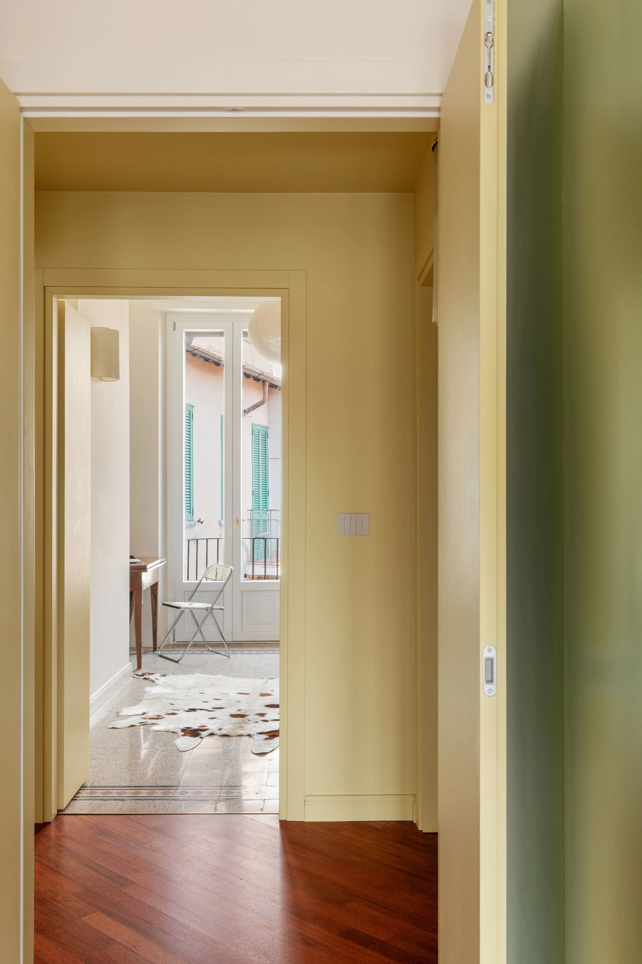 Vista di un ingresso con pavimento in legno, porte di passaggio e una porta finestra che dà su un balcone con tapparella verde e edificio esterno visibile.