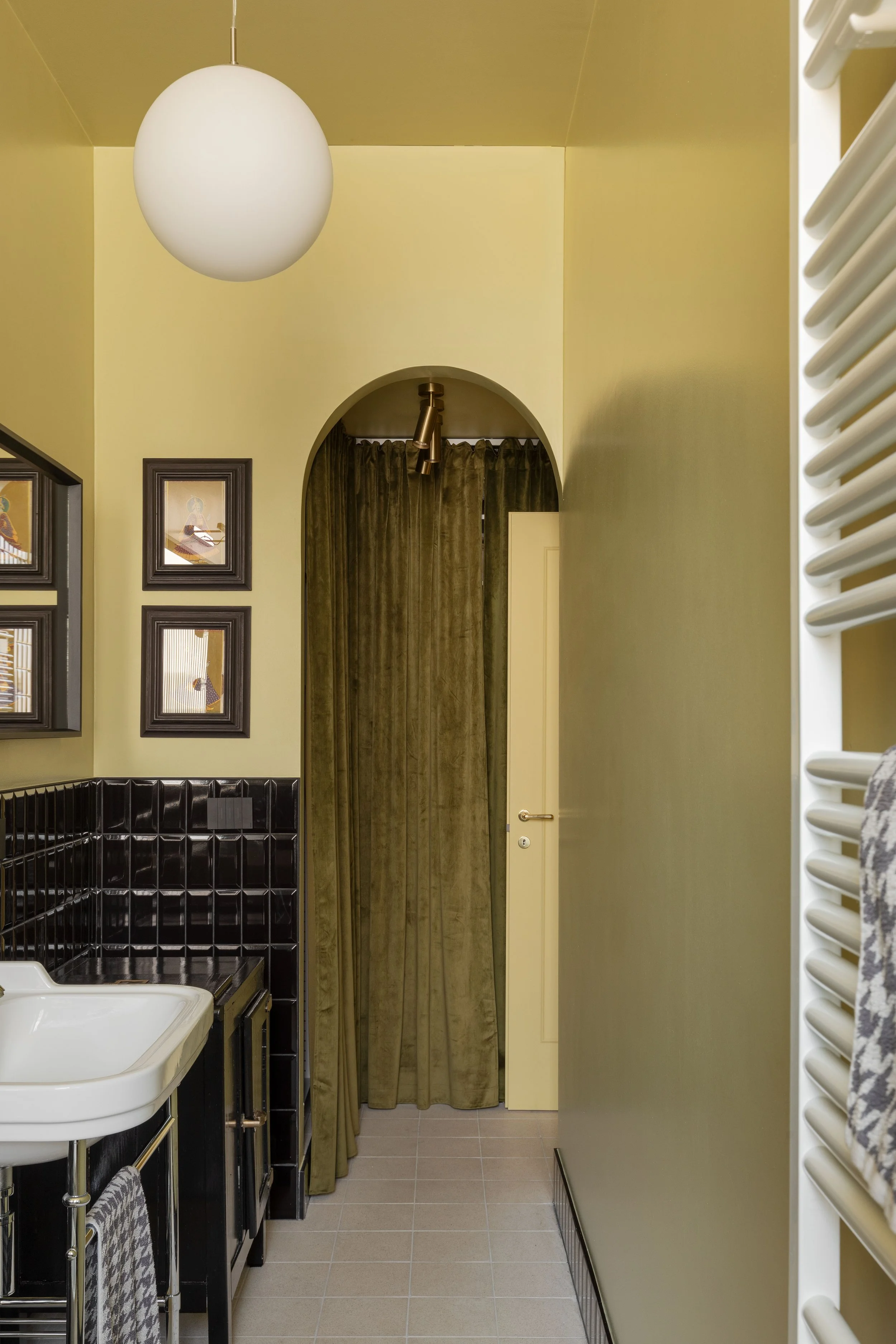 Interno di una cucina con pareti beige, lampada rotonda bianca, quadri alle pareti, presenza di forno e lavandino, tenda verde scuro e termosifoni bianchi.