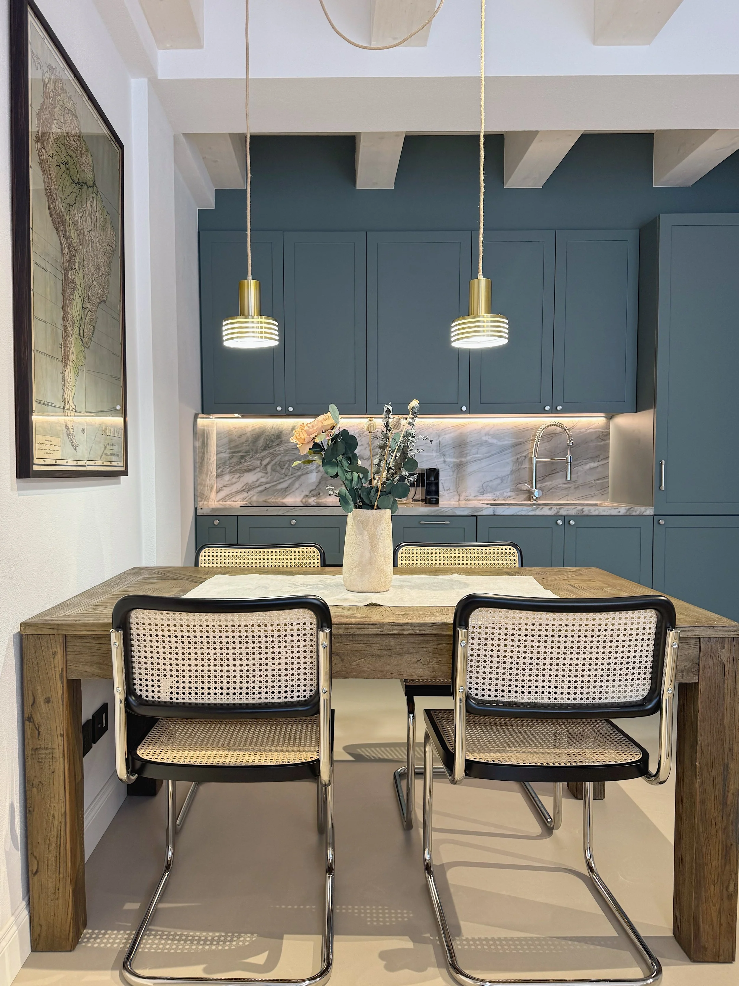 Camera da pranzo con tavolo in legno, quattro sedie con struttura in metallo e seduta in cannetté, al centro un vaso con fiori. Sullo sfondo una cucina con armadietti blu, piano e schienale in marmo beige e illuminazione a LED.