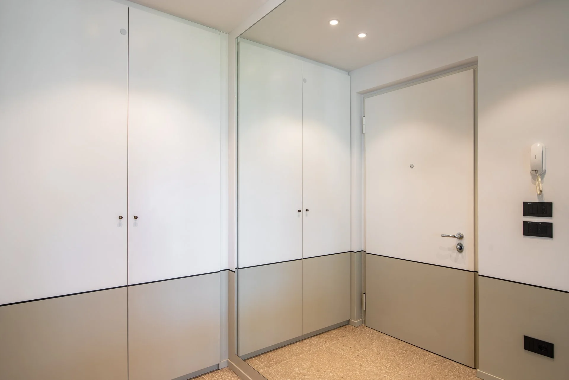 Entrata di un appartamento con pareti bianche e armadiature con ante bianche e maniglie nere, una porta bianca con maniglia metallica, e un interfono bianco sulla parete a destra.