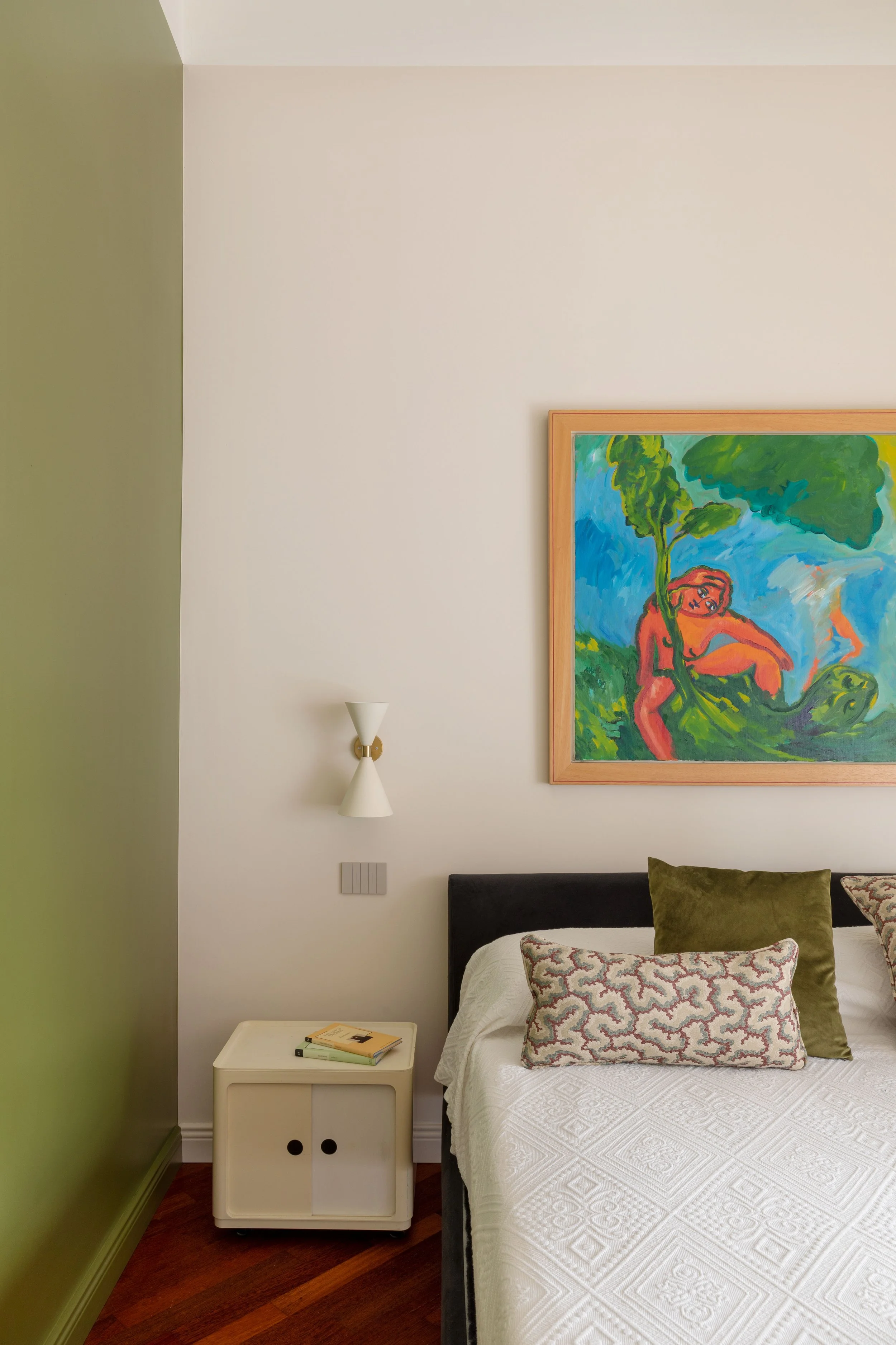 Una camera da letto con pareti bianche e una parete verde. Sopra il letto c'è un quadro con un dipinto colorato. Alla sinistra del letto una lampada da muro e un comodino bianco con alcuni libri. Il letto ha cuscini decorativi, uno verde e uno con un