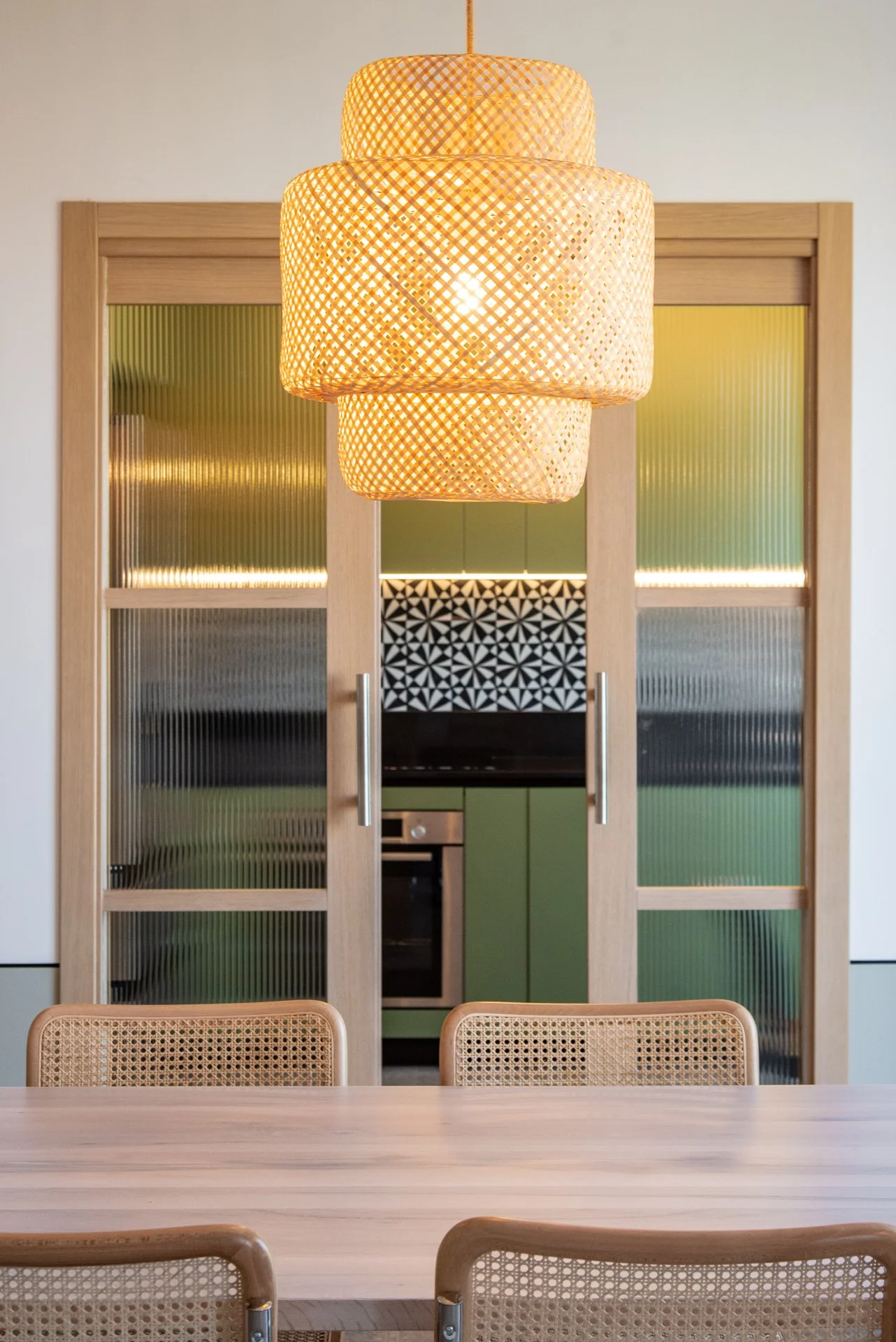 Lampadario di paglia sopra un tavolo di legno e sedie in bambù, con cucina moderna sullo sfondo.