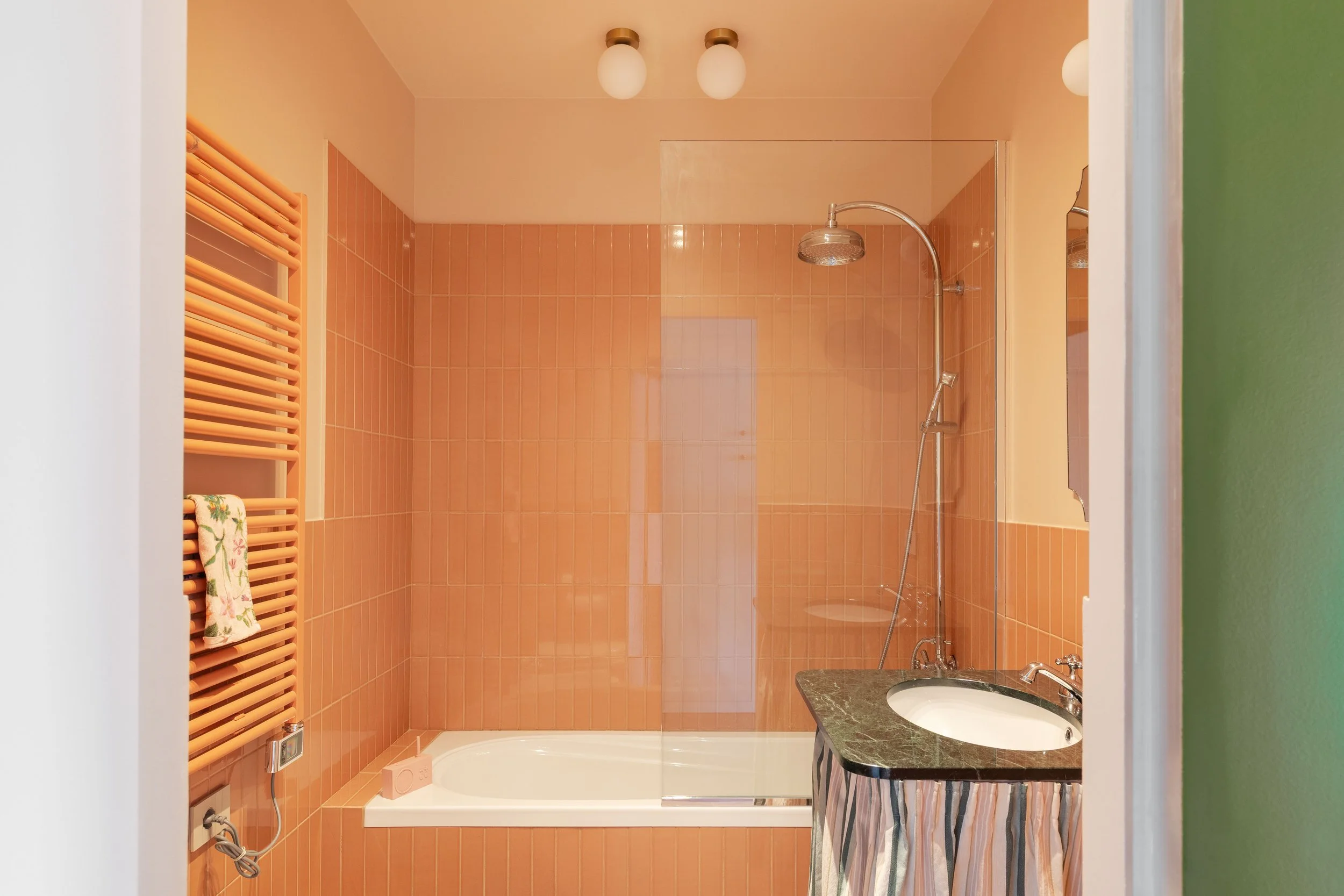 Bagno con pareti rivestite di piastrelle arancioni, doccia con soffione a pioggia, lavabo con mobile e specchio, scaldasalviette a rulli e finestra.