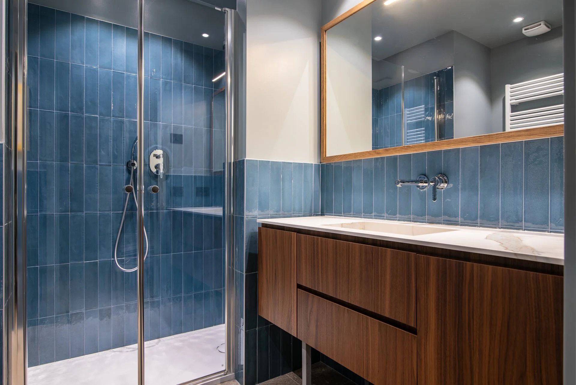 Bagno moderno con pareti rivestite di piastrelle blu, lavabo in marmo su mobile in legno, grande specchio con cornice in legno, doccia con porte in vetro e soffione a soffitto.