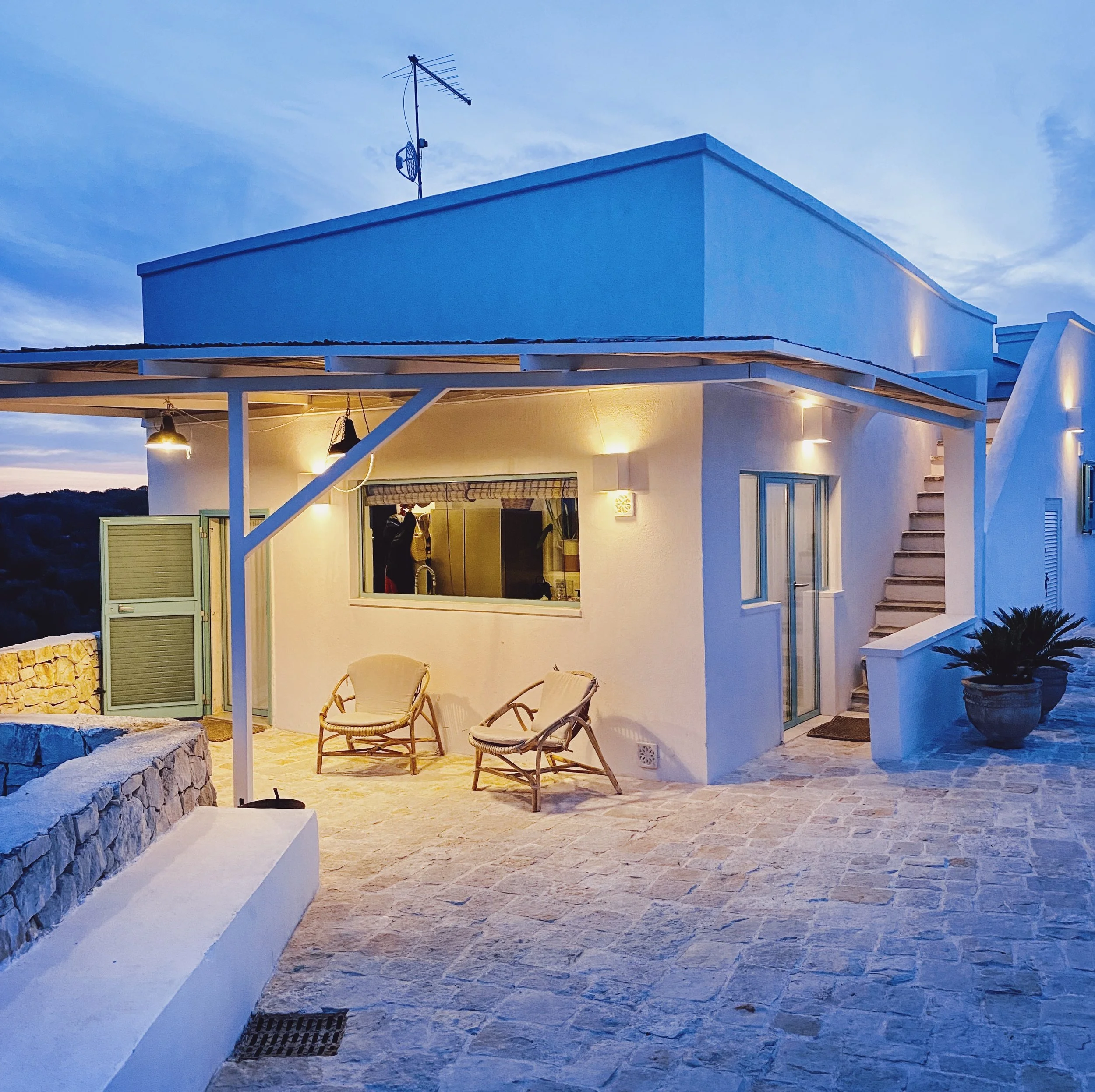 Casa bianca con sedia e panca in pietra, piante in vaso, illuminazione esterna, vista al tramonto
