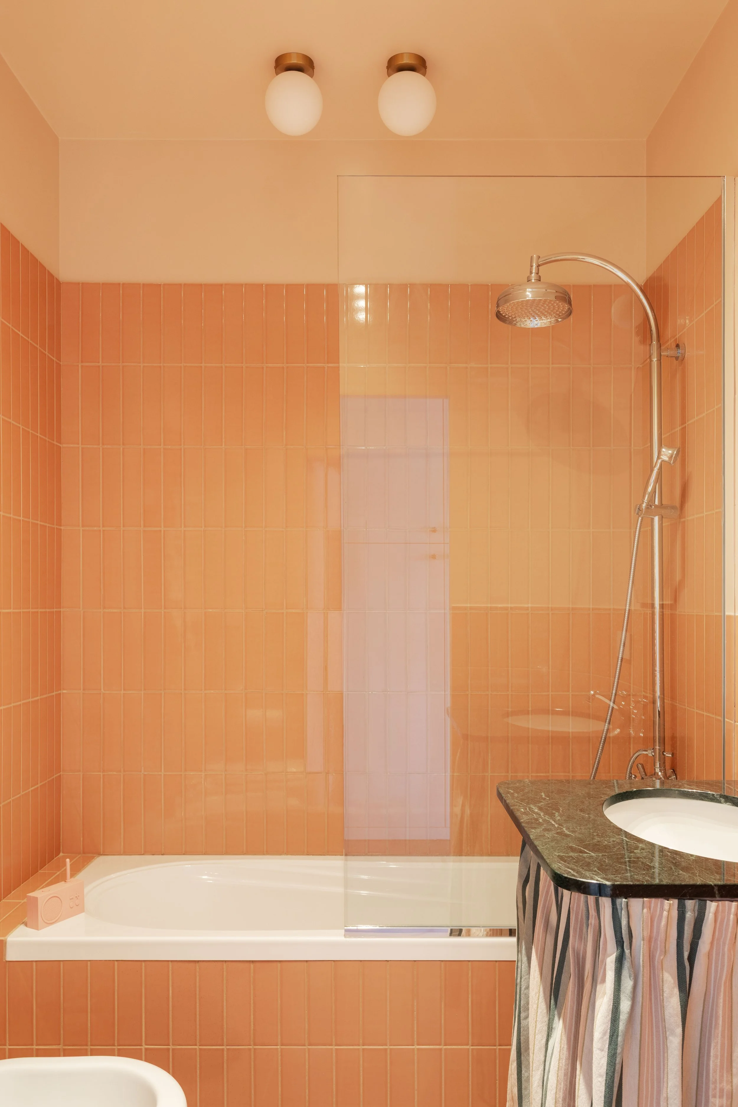 Stanza da bagno con pareti in piastrelle beige e arancioni, soffitto con due plafoniere, vasca da bagno, soffione doccia e lavabo con mobile sotto con tende a righe.