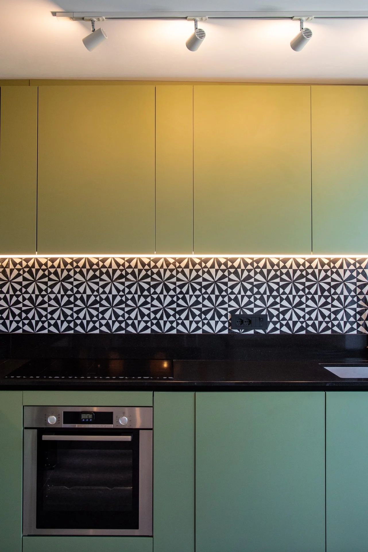 Cucina moderna con pensili verdi menta, piano di lavoro nero, piastrelle decorative nere e bianche con motivo geometrico, uscita USB sulla parete e forno elettrico in acciaio inox.