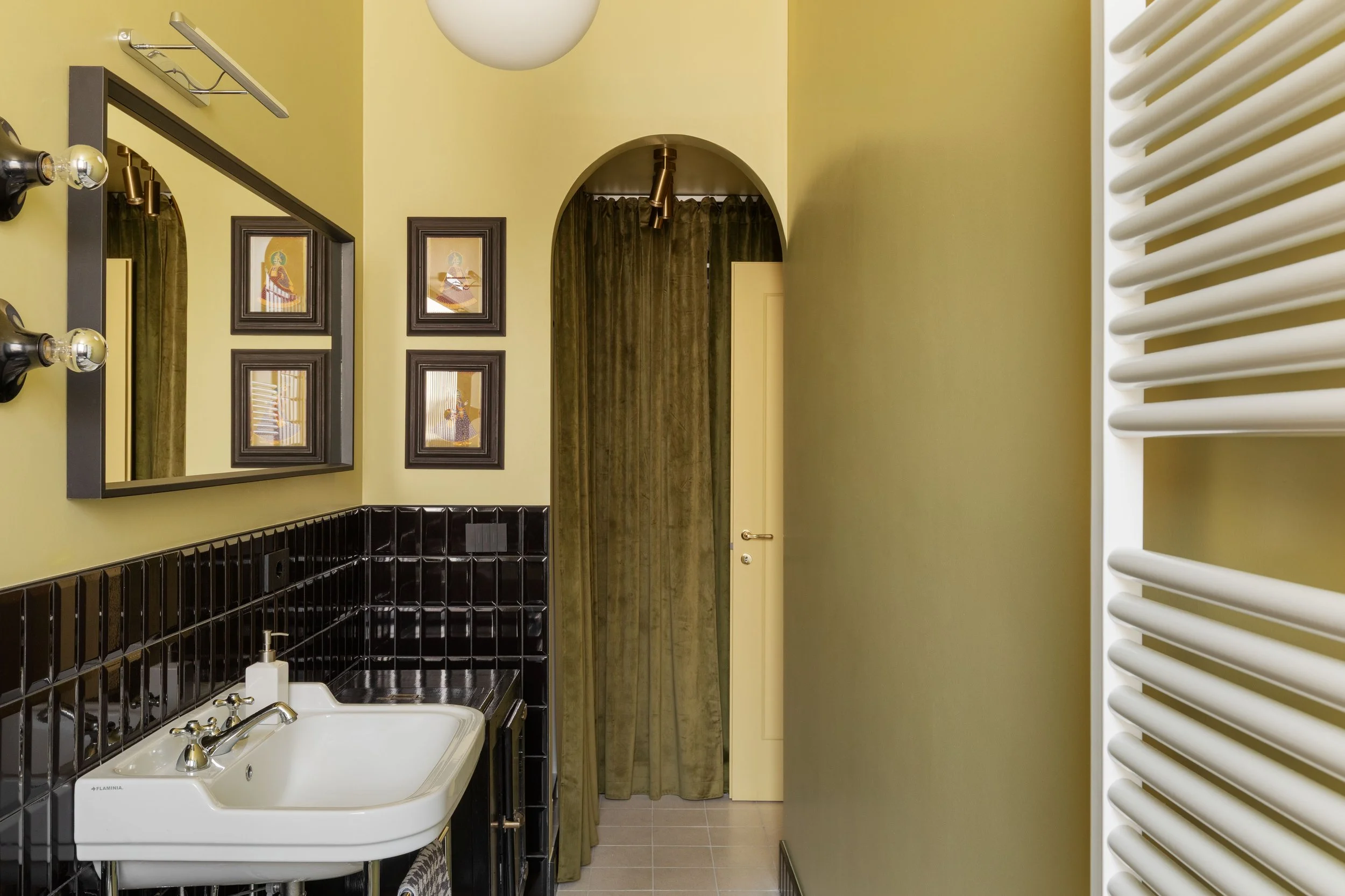 Interno di un bagno con pareti gialle, sembianze vintage, con quadri alle pareti, uno specchio e un lavandino con rubinetti in primo piano, una tenda in velluto e una porta in fondo.