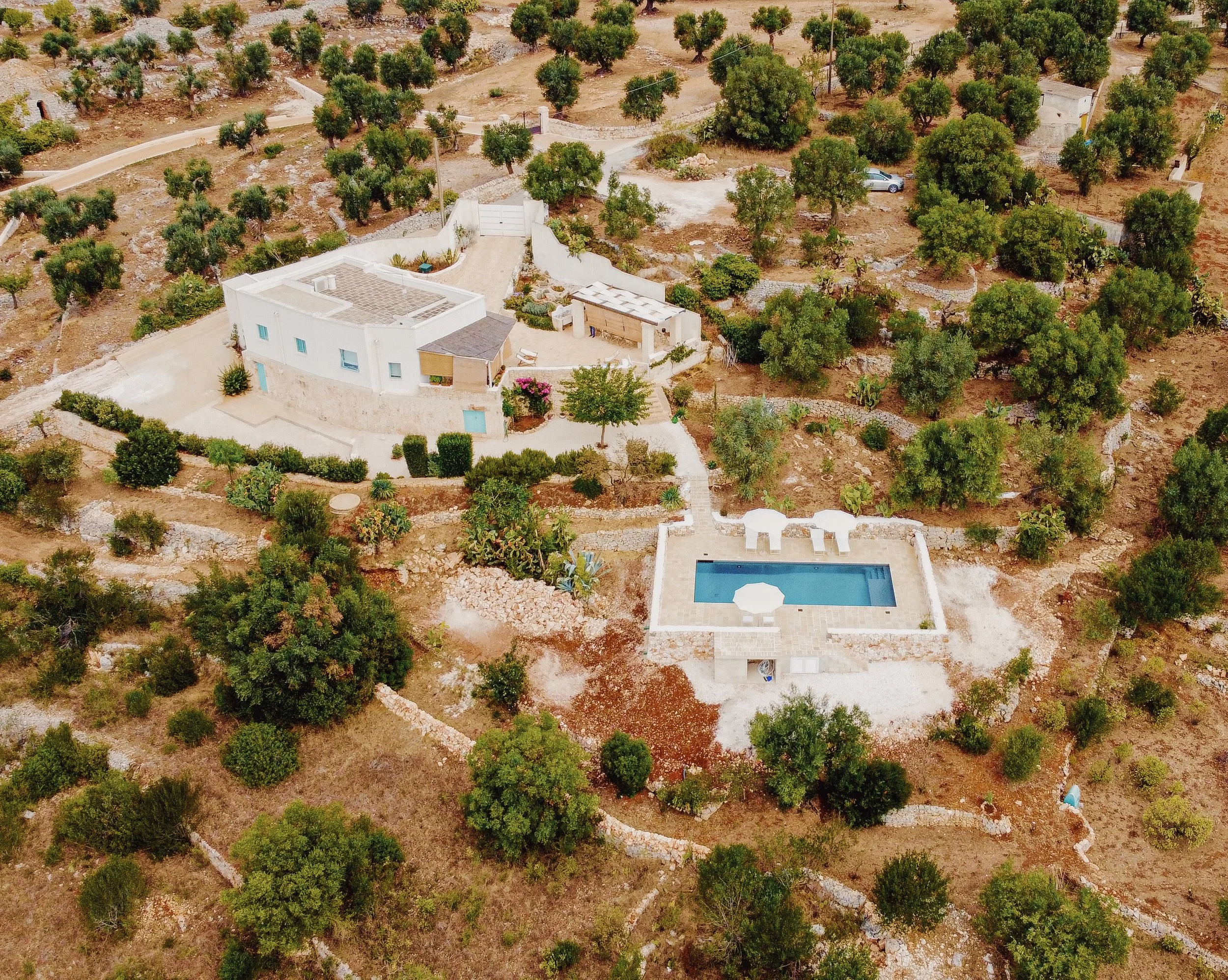 Vista aerea di una casa bianca con piscine e giardino in un ambiente rurale, con molti alberi e terreni aridi circostanti.