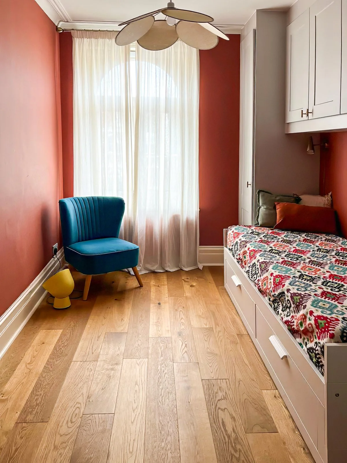 Stanza con pareti color terracotta, un grande finestrone con tende leggere, un letto con coperta colorata, un armadio bianco e una sedia elegante blu, lampada decorativa sopra il letto.