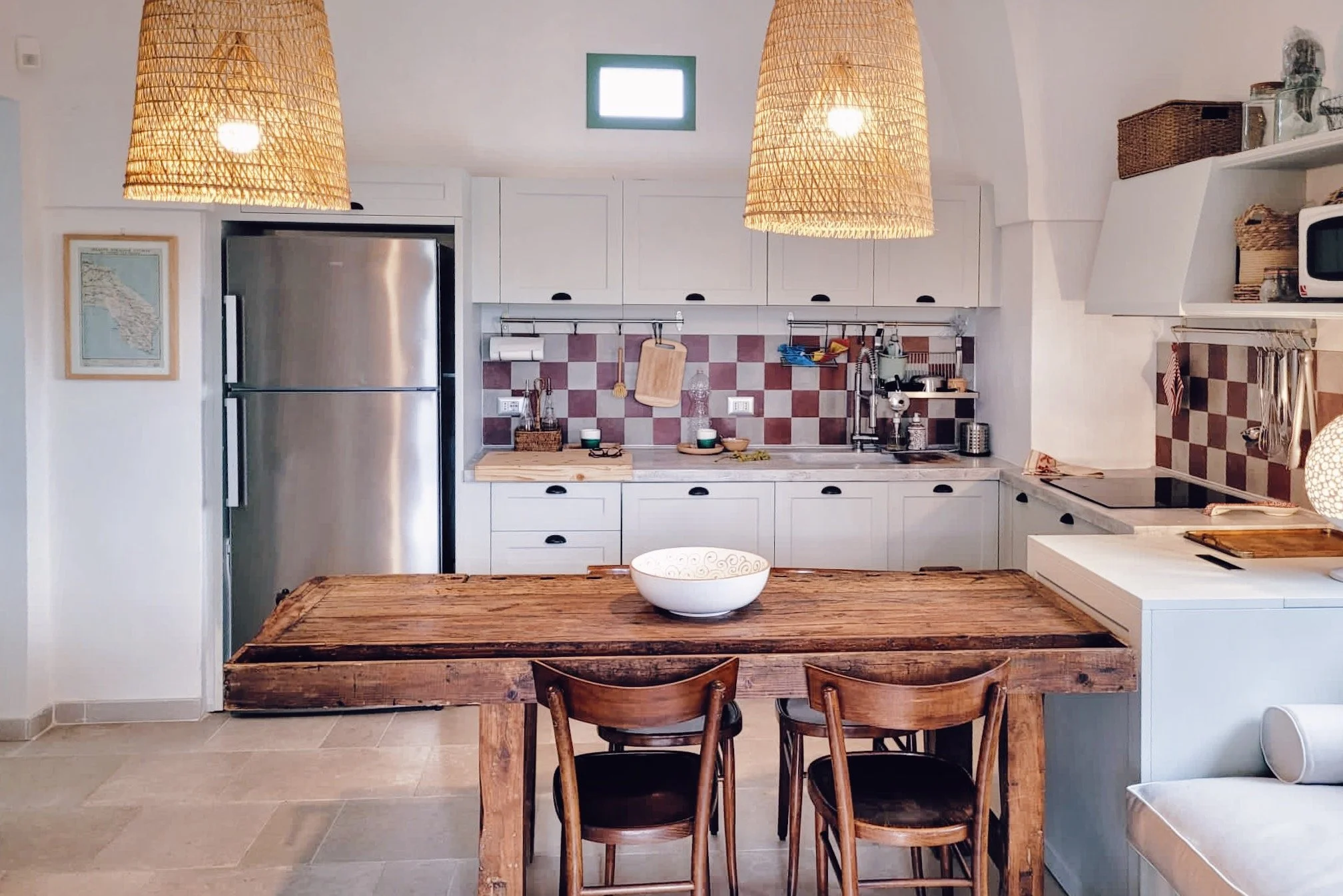 Cucina moderna con mobili bianchi, un frigorifero in acciaio, un tavolo in legno massiccio, luci a sospensione di vimini, una parete con piastrelle a scacchi rossi e beige, e vari accessori da cucina.