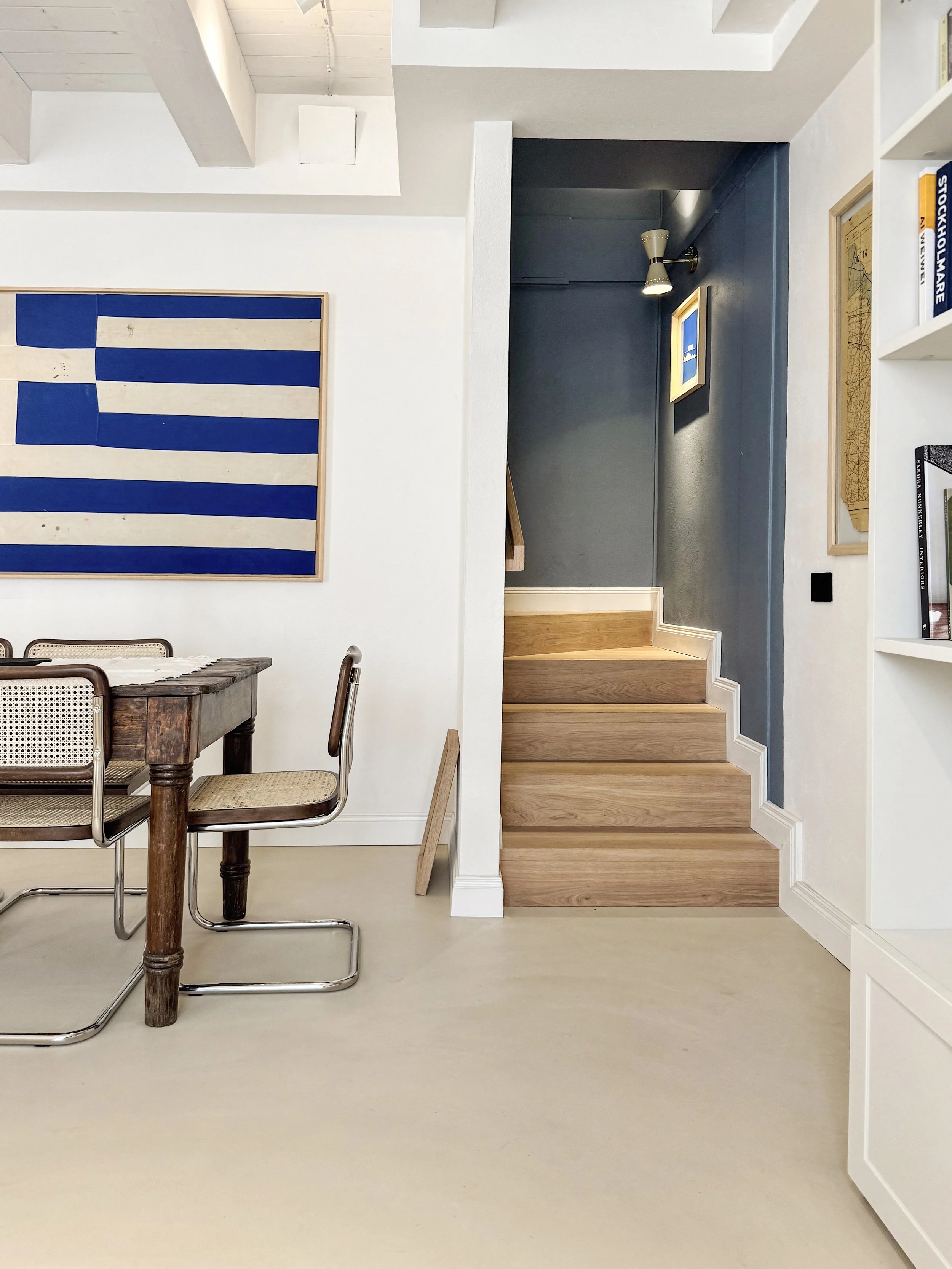 Interno di una casa con pareti bianche, una scala di legno, una cornice con bandiera blu e bianca, una libreria con libri e decorazioni, una lampada a parete e un tavolo rustico con sedie