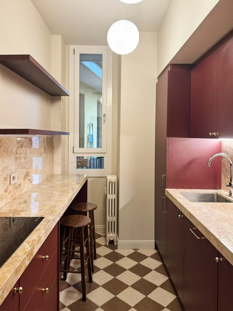 Una cucina compatta con armadietti bordeaux, un bancone in marmo beige, due sgabelli di legno, due lampade a sfera bianche e una finestra che dà su un cortile con un'attrezzatura da esterno.