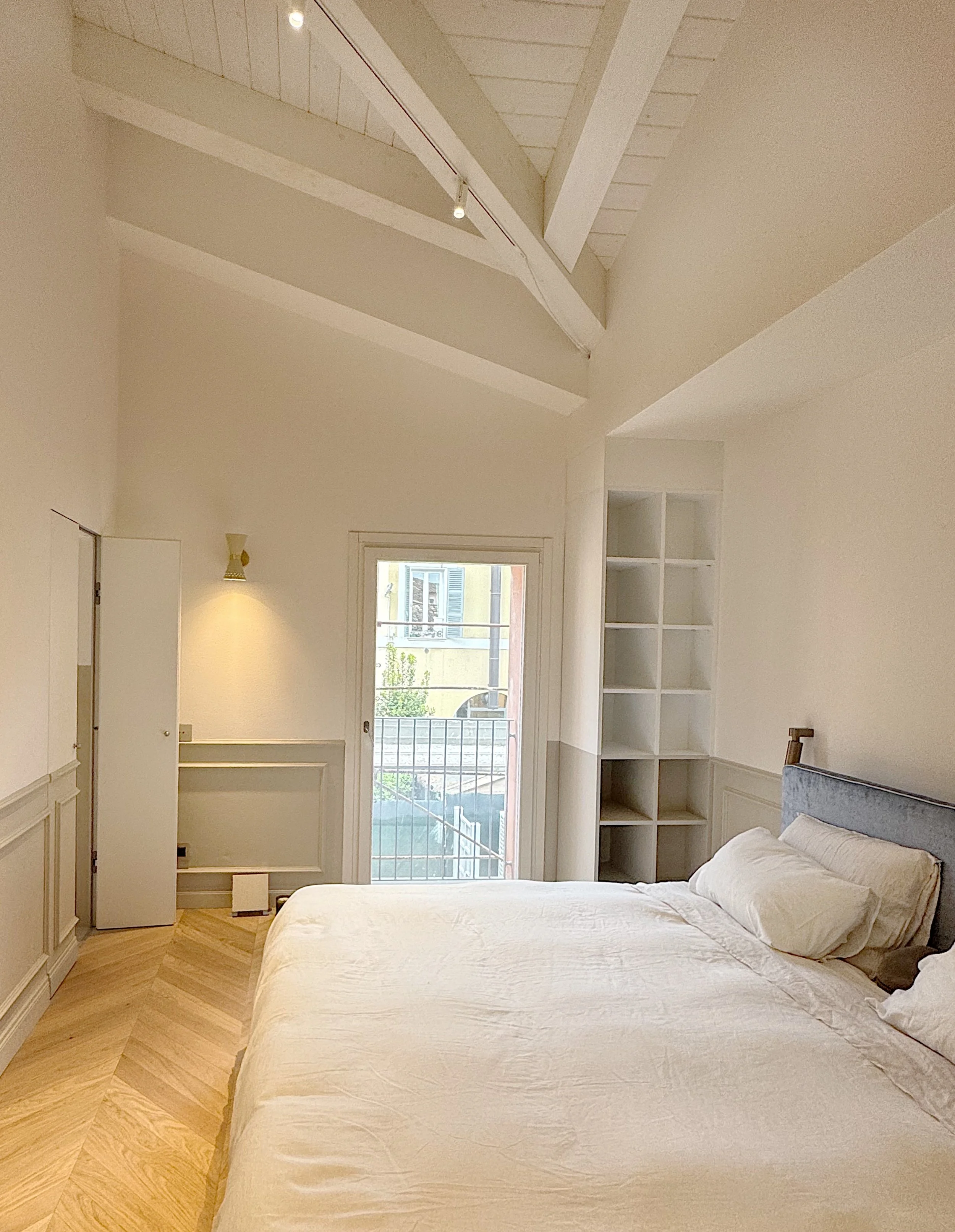 Camera da letto con letto matrimoniale con lenzuola bianche, cuscini e copriletto, pareti chiare, armadiature e librerie bianche, finestra con vista su un balcone e un edificio vicino, luce naturale e lampada acceso sulla parete.