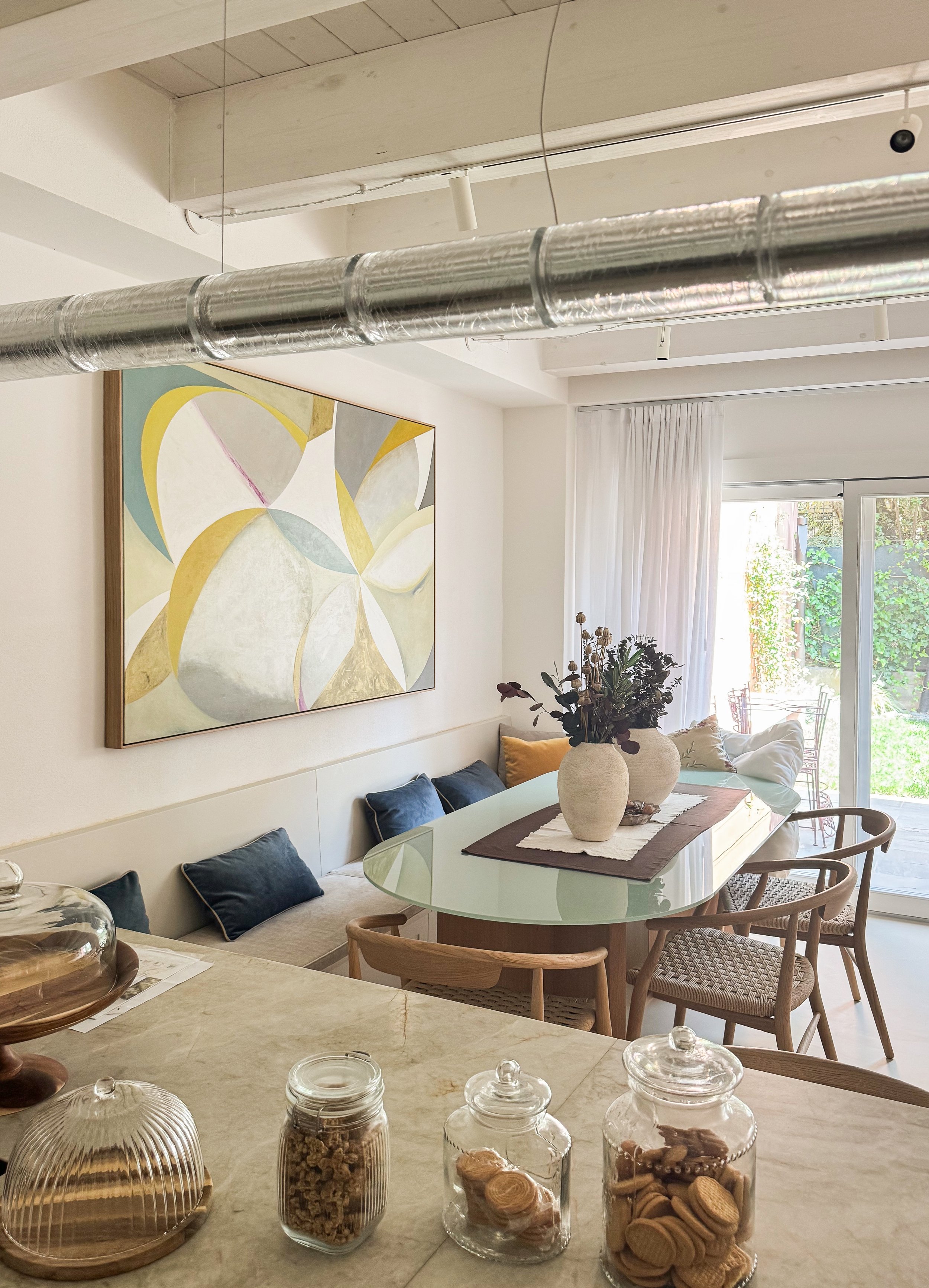 Interno di una zona pranzo con un tavolo rotondo in vetro trasparente, decorato con vasi di vari tipi di biscotti e dolci in barattoli di vetro trasparenti. Sullo sfondo, pareti con armadio a muro, un quadro astratto appeso alla parete, e una grande 