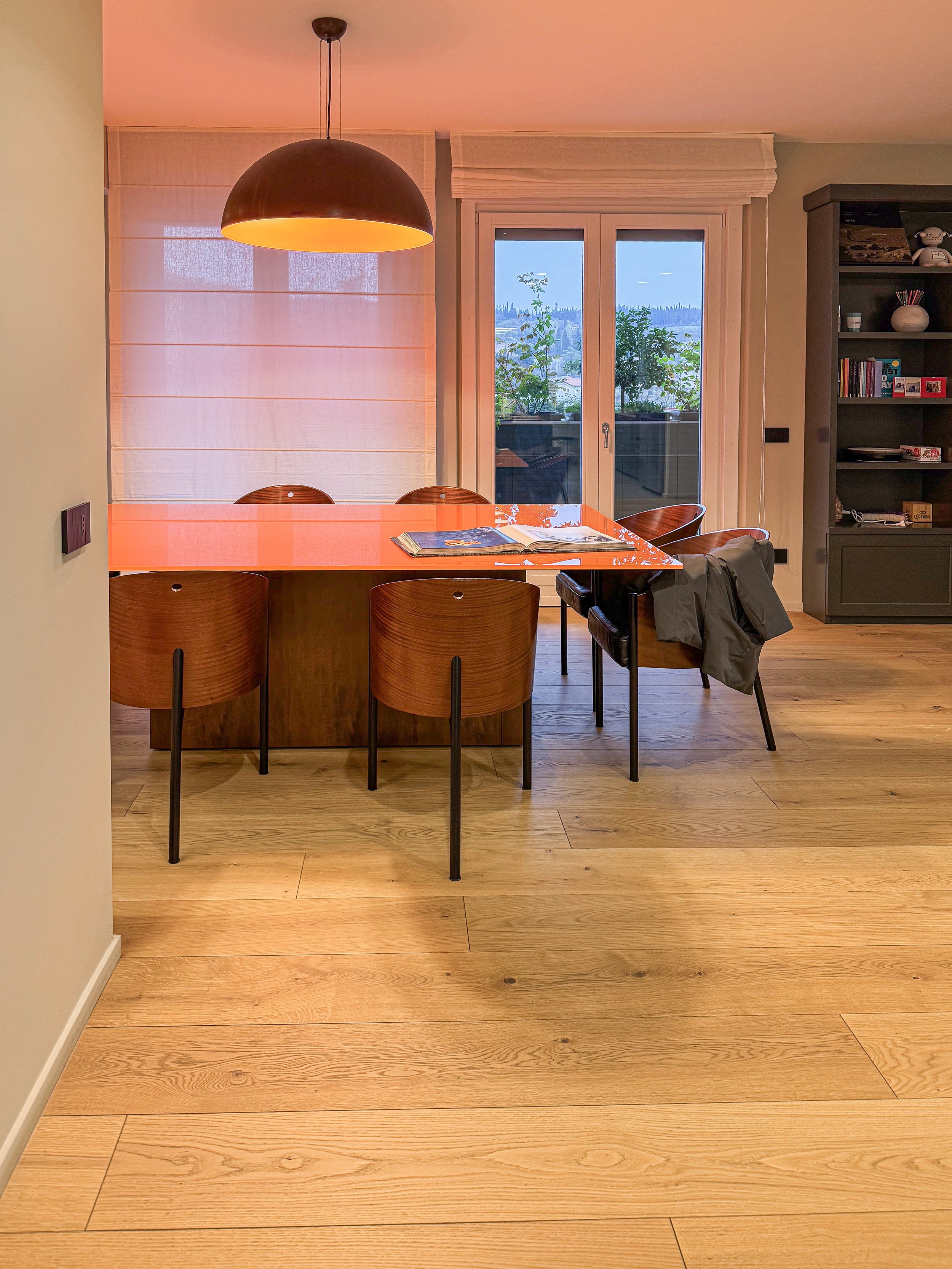 Sala da pranzo con tavolo rosso e sedie di legno, lampada a soffitto nera, finestre con vista su balcone e natura, libreria a muro grigia con libri e decorazioni.