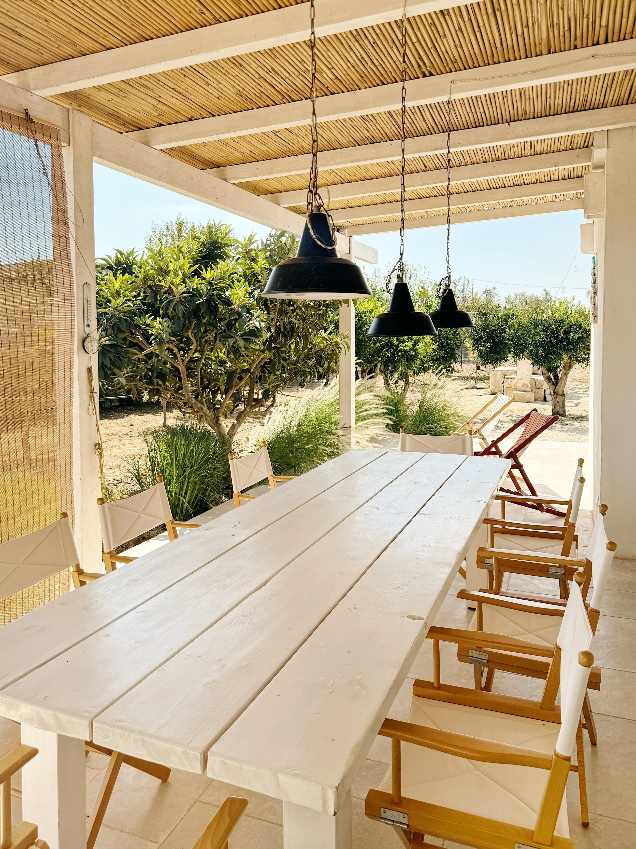 Zona pranzo coperta con tavolo bianco in legno, sedie di legno con seduta in tessuto bianco, e lampade nere sospese sopra il tavolo. Sullo sfondo, piante e alberi in un giardino soleggiato.