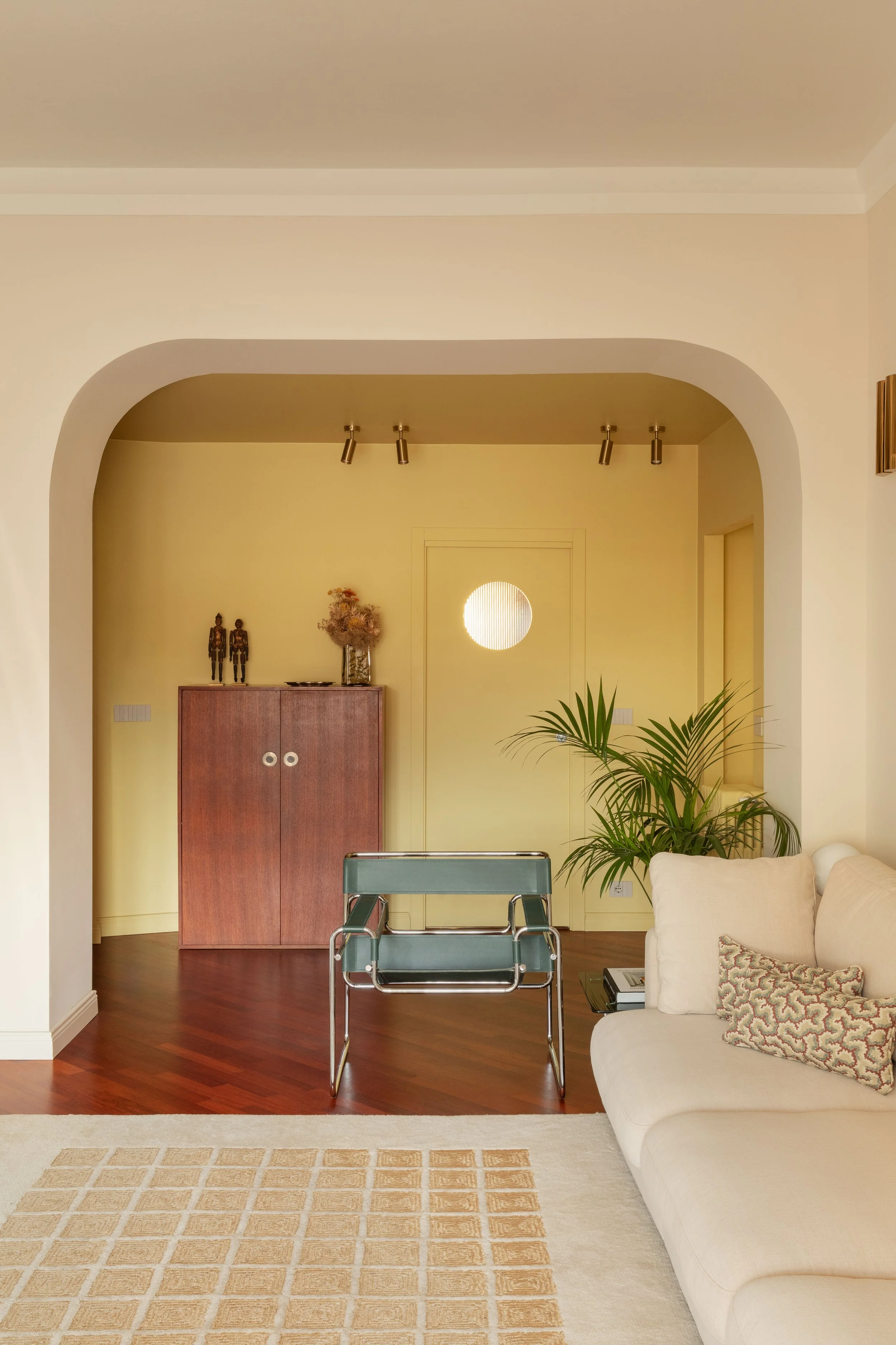 Intero soggiorno con pareti gialle, arreda con armadietto in legno, divano beige, poltrona in metallo con sedile in pelle blu, tappeto beige con decorativi quadrati e piante verdi, illuminazione a soffitto con faretti.