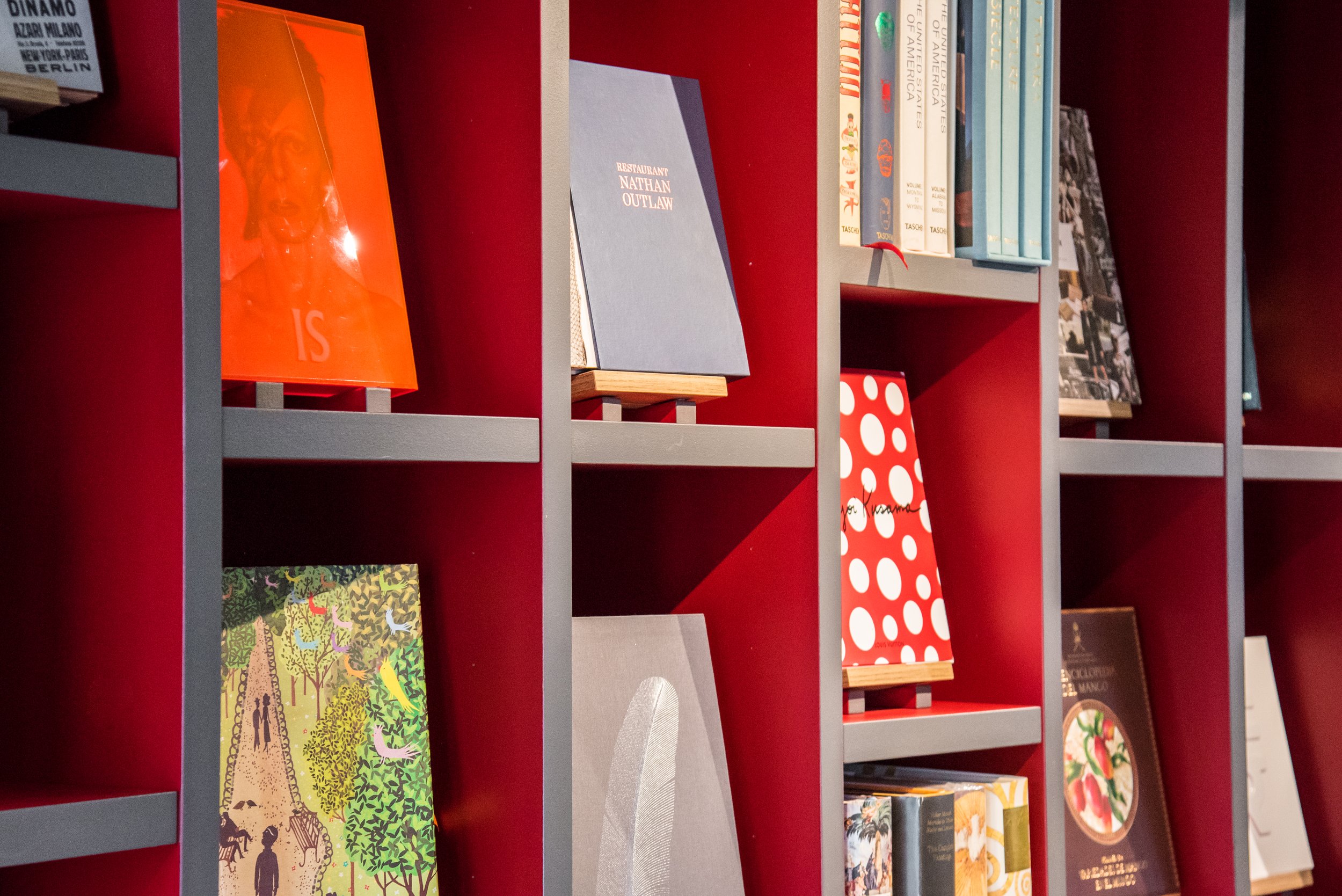 Mensola rossa con diversi libri di varia grandezza e colore, alcuni aperti e altri impilati, su uno sfondo grigio e rosso.