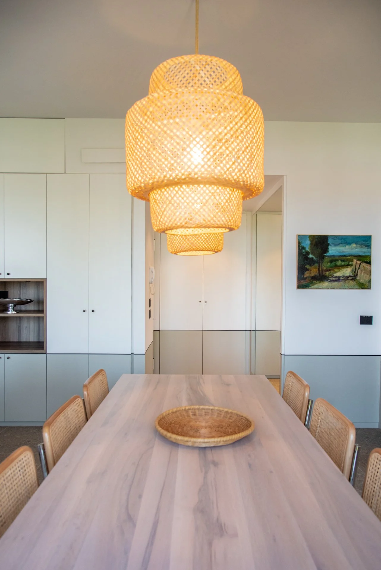 L'immagine mostra una sala da pranzo moderna con un lungo tavolo di legno chiaro e sei sedie con sedili di paglia. Sopra il tavolo pendono tre lampadari di vimini di forma cilindrica.
