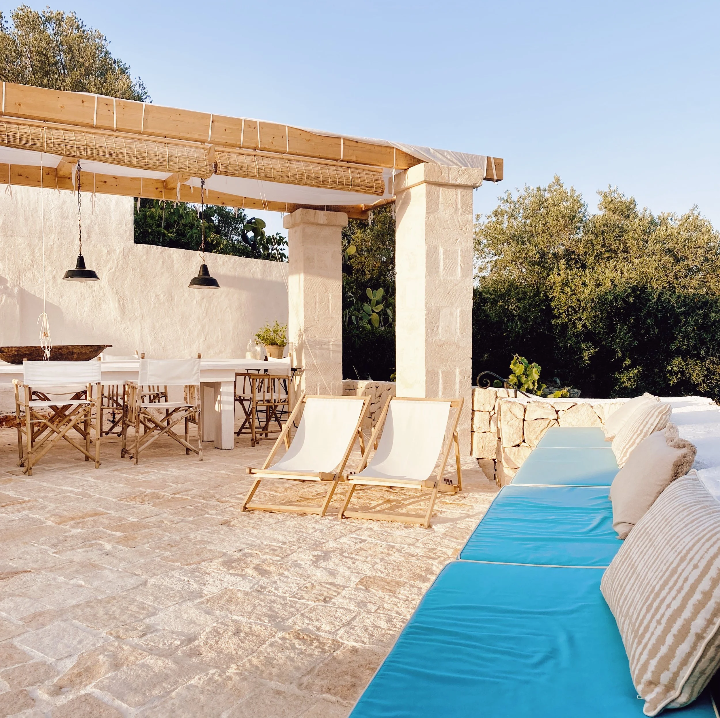Terrazza all'aperto con sedie, lettini e divano con cuscini, sotto una tettoia di legno, con sfondo di alberi e cielo azzurro.
